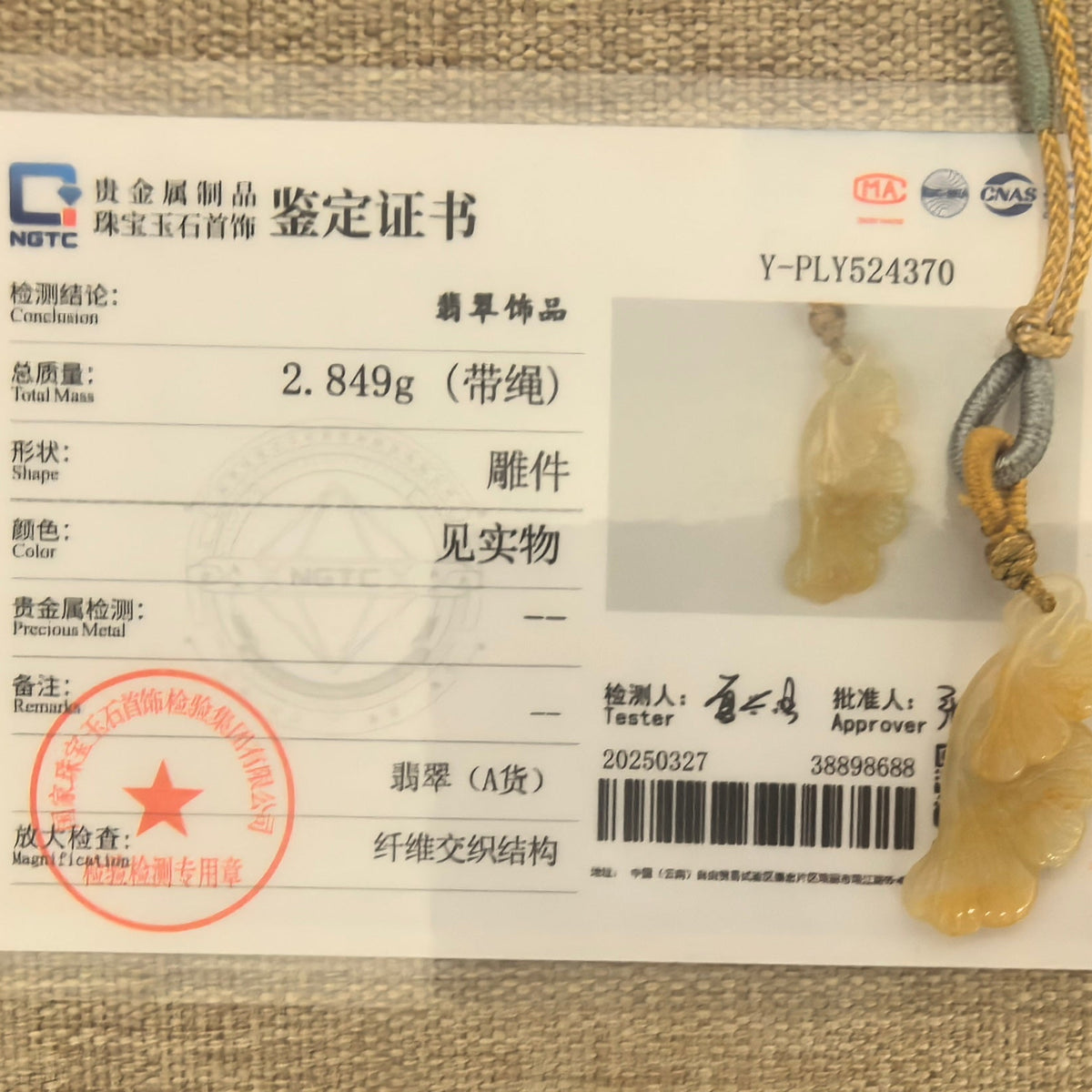 Yellow jadeite ginkgo leaf pendant beside its matching NGTC certificate on a woven background.