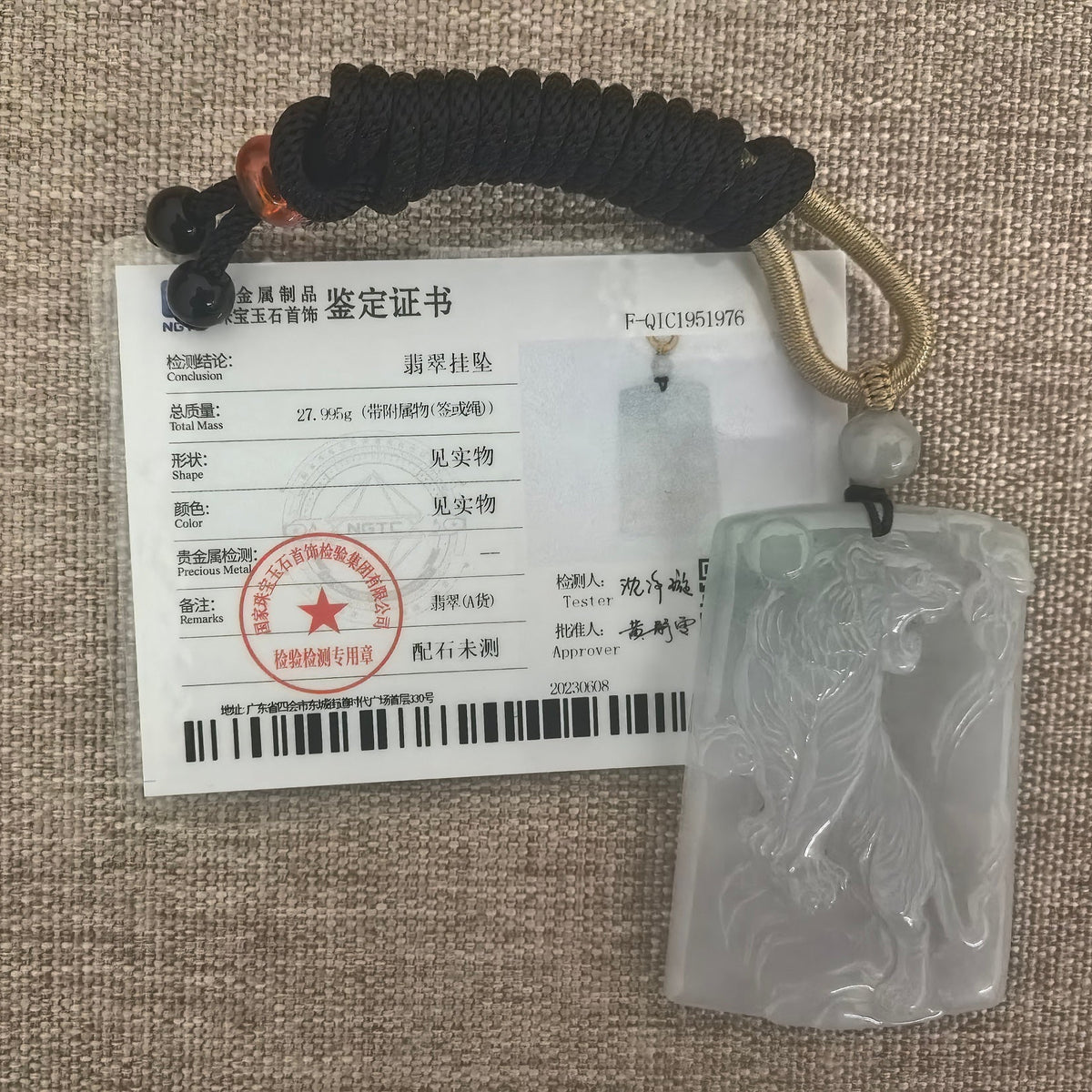 Pale green jadeite tiger plaque pendant with a knotted cord beside its matching NGTC certificate on a woven background.