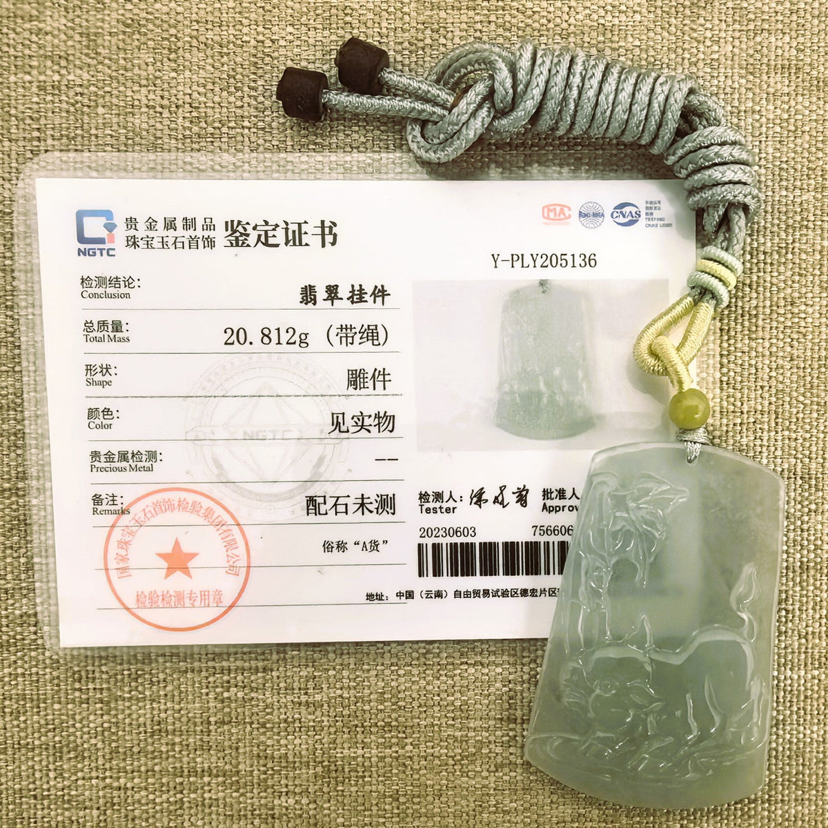 Pale green jadeite Pig Plaque Pendant with a knotted cord and yellow accent bead beside its matching NGTC certificate on a woven background.