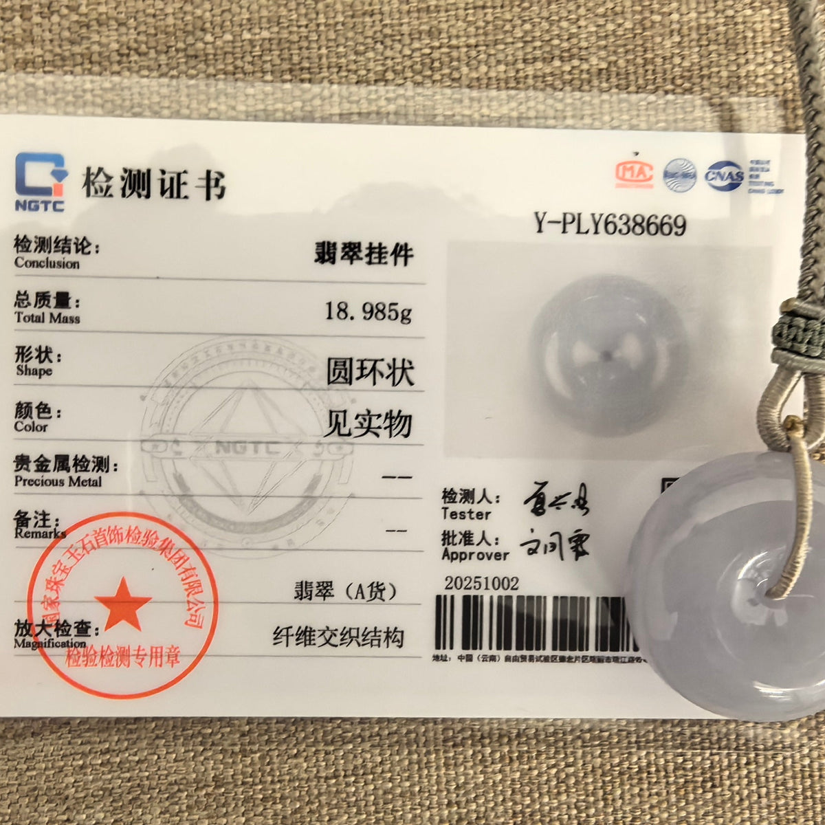 Lavender jadeite Peace Pendant with a gray braided cord beside its matching NGTC certificate on a woven background.