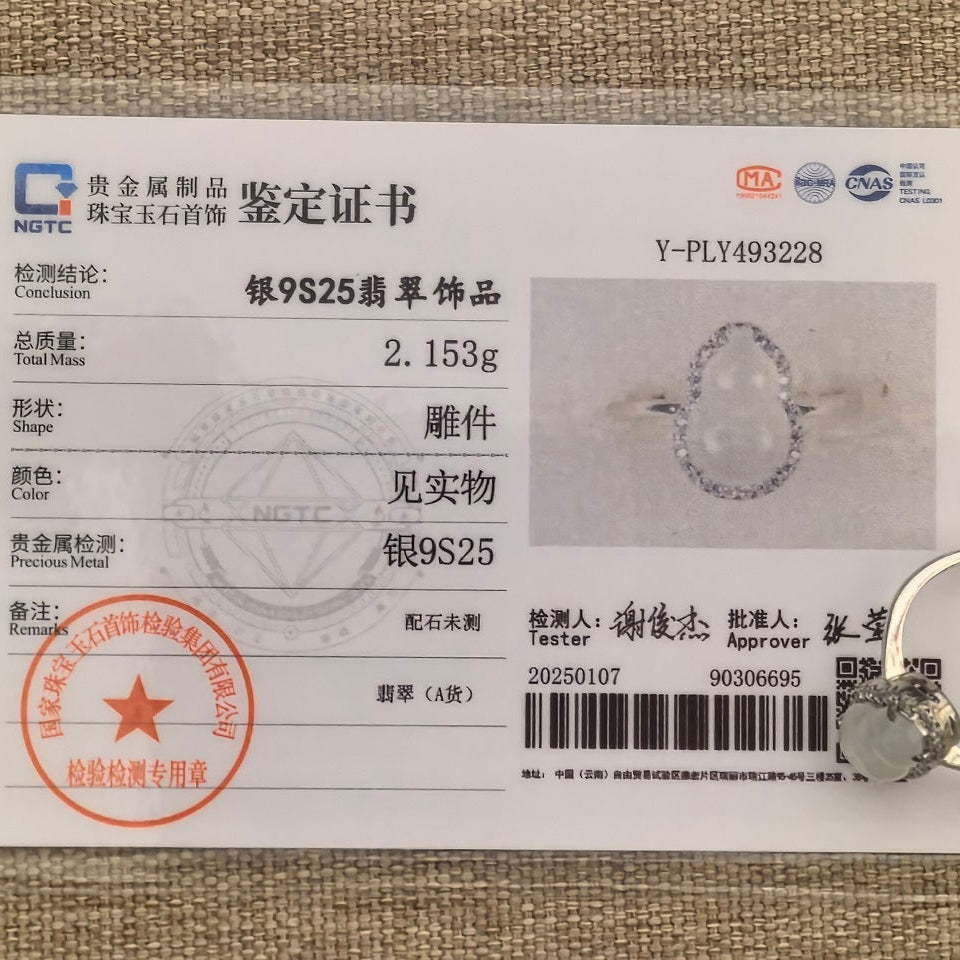 White jadeite gourd ring beside its matching NGTC certificate on woven fabric, with the silver setting and ring band visible.