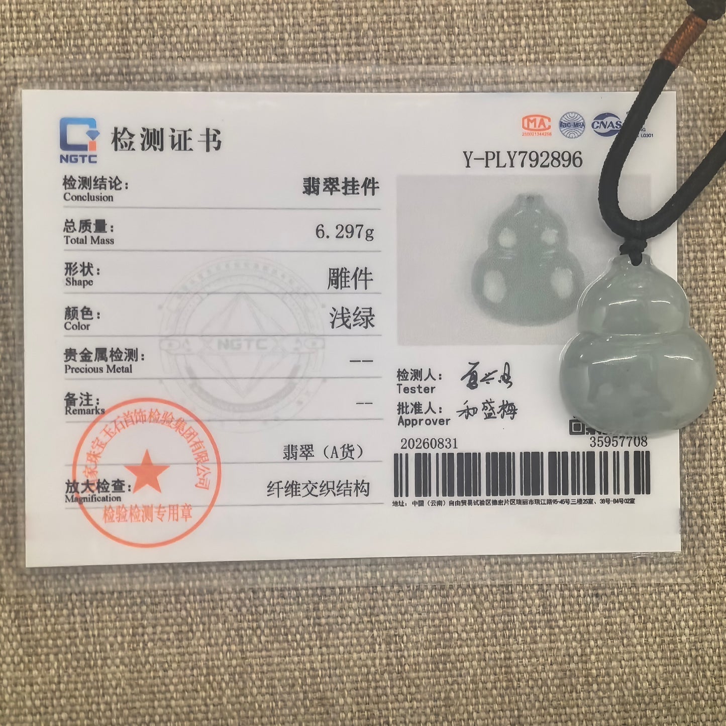 Light green jadeite gourd pendant and black cord displayed with the matching NGTC certificate on woven fabric.