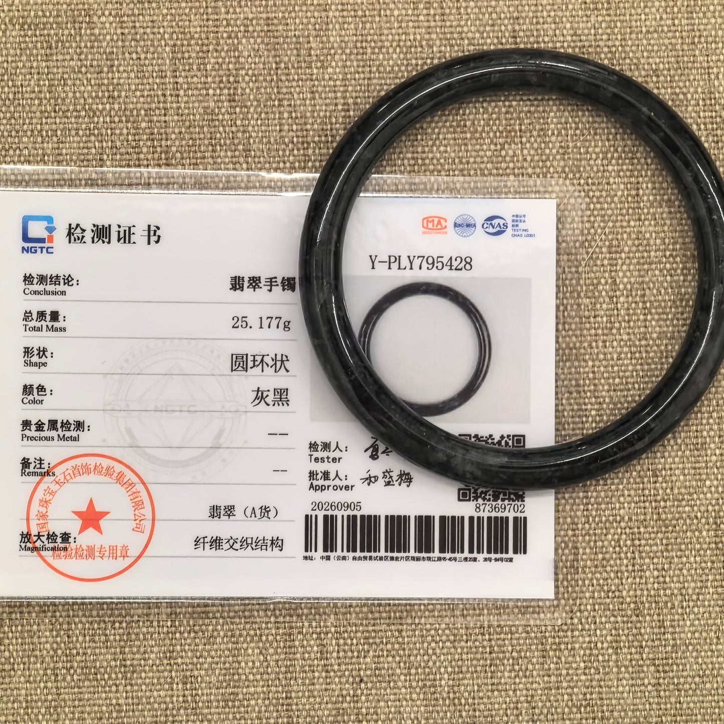 Black and gray jadeite bangle beside its matching NGTC certificate on tan woven fabric.