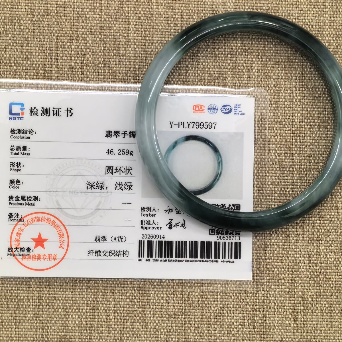 Light and dark green jadeite bangle displayed with its matching NGTC certificate on woven fabric.