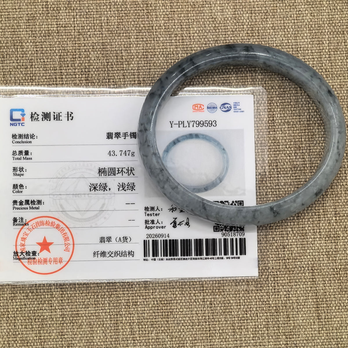 Blue floating pattern oval jadeite bangle displayed with its matching NGTC certificate on a woven background.