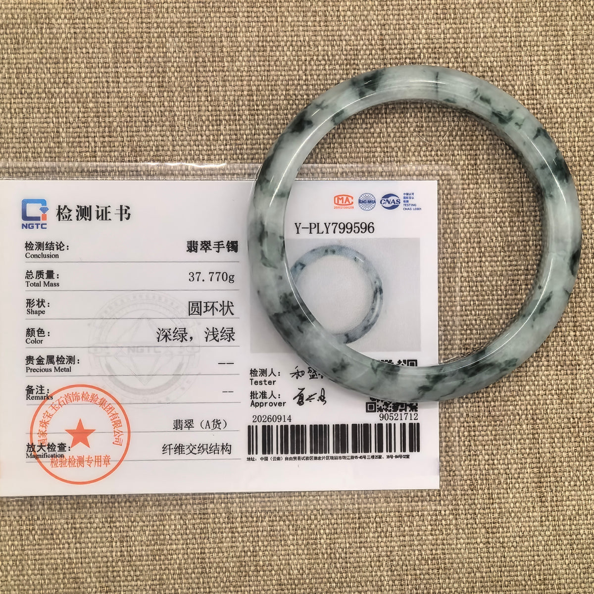 Green floating flower jadeite bangle displayed with its matching NGTC certificate on a tan woven background.