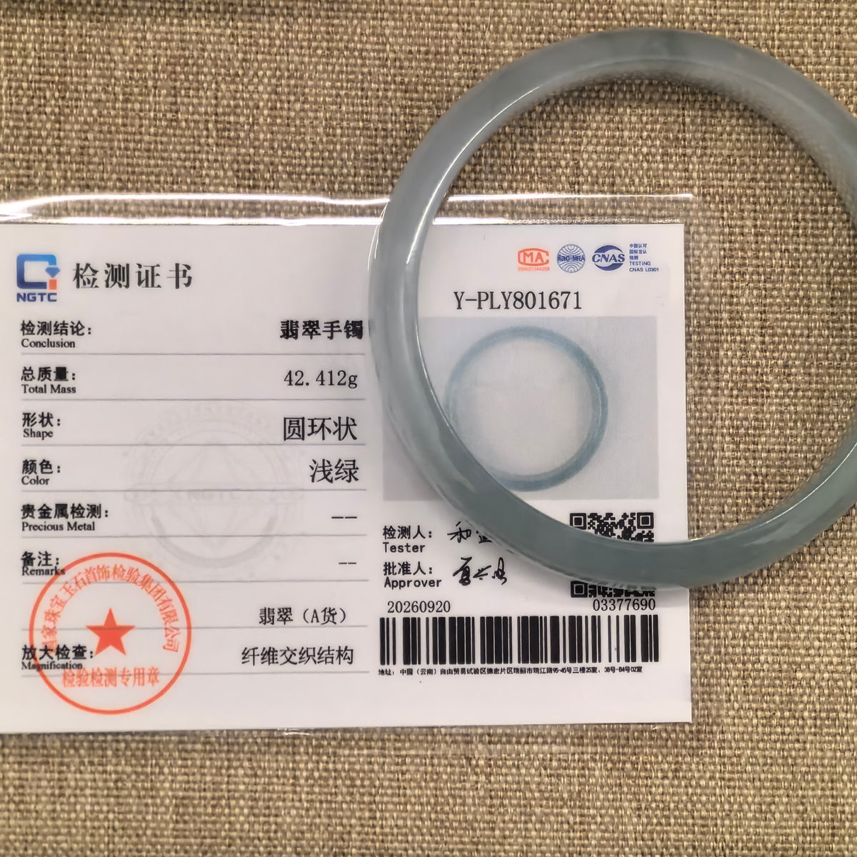 Pale green jadeite bangle displayed with its matching NGTC certificate on a tan woven background.