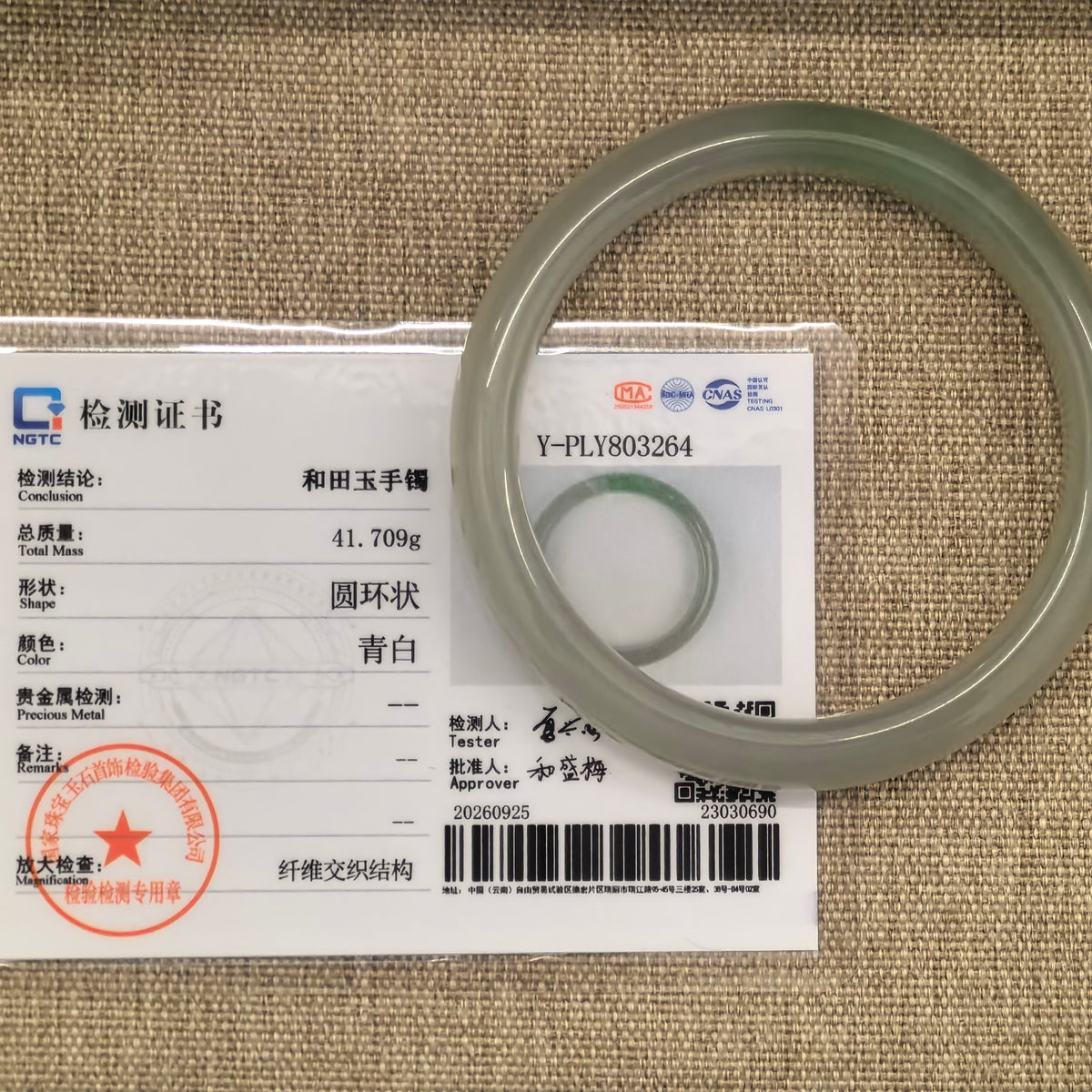 Greenish white Hetian jade bangle with a green accent displayed alongside its matching NGTC certificate on a woven background.