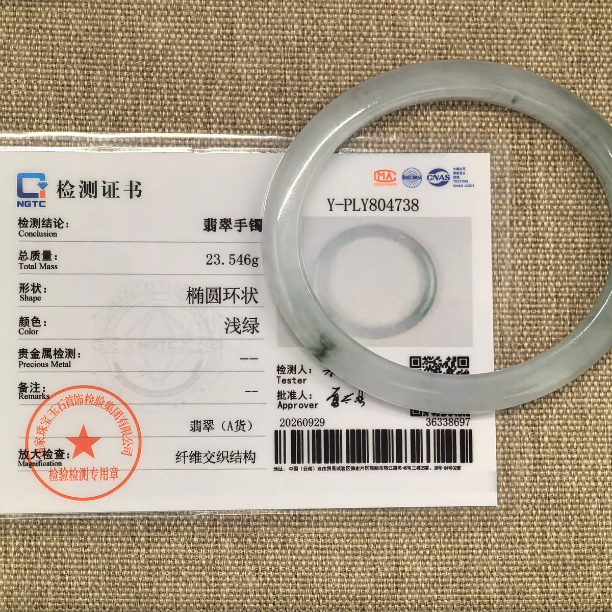 Pale green oval jadeite bangle displayed over its matching NGTC certificate on a woven background.
