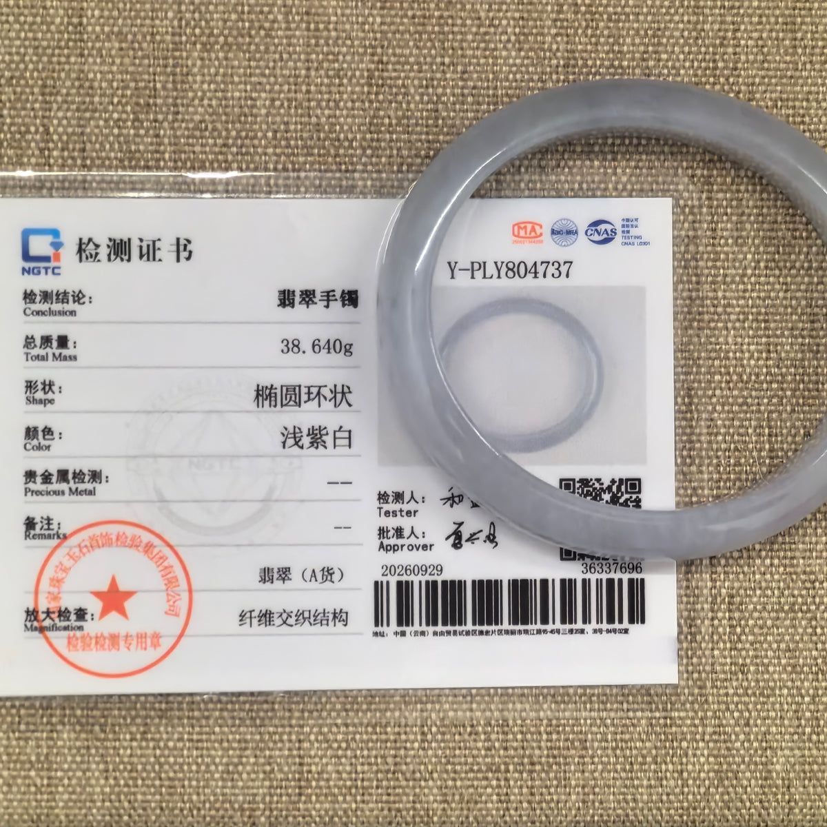 Pale lavender oval jadeite bangle displayed over its matching NGTC Grade-A jadeite certificate on a woven background.