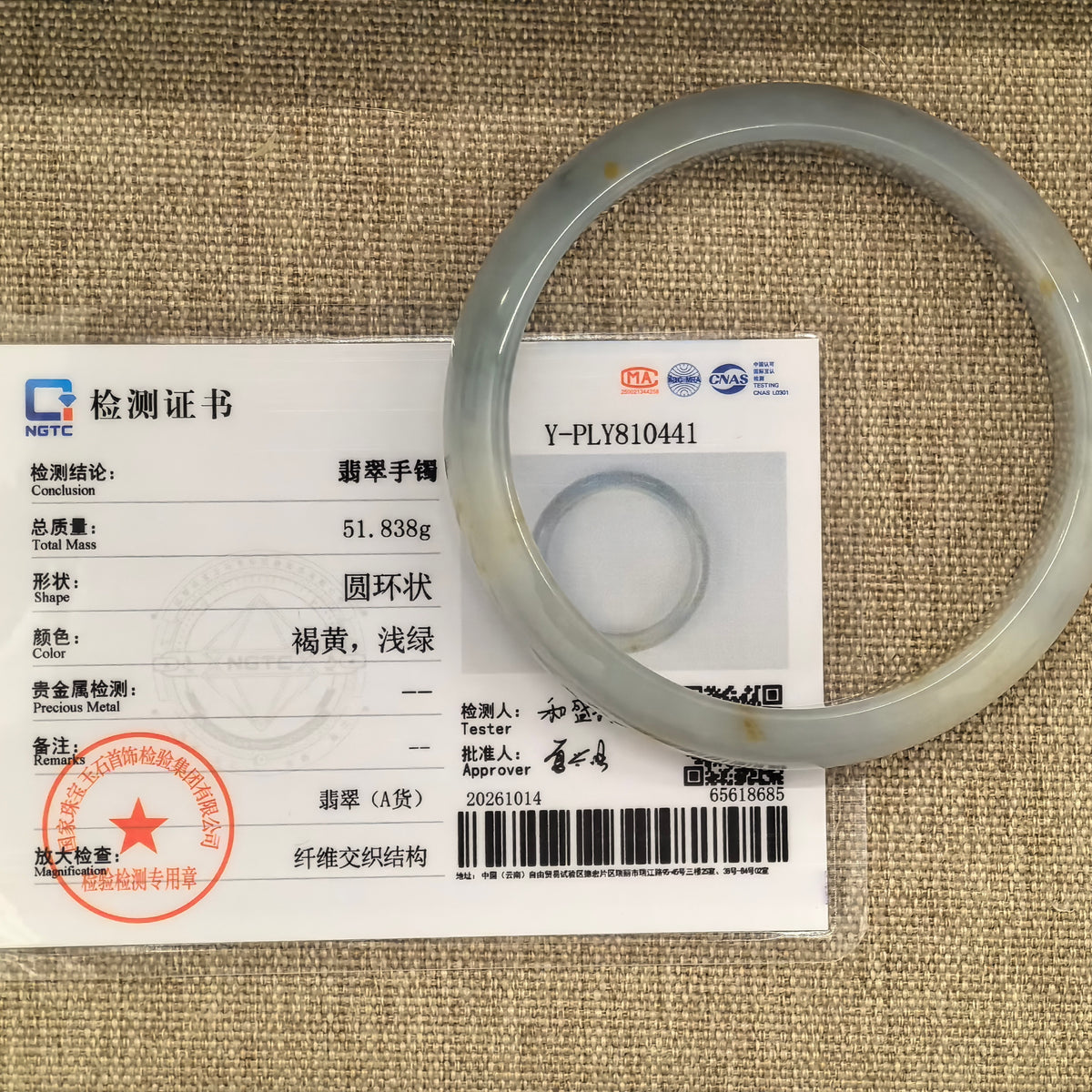 White and pale green jadeite bangle with golden-brown flecks beside its matching NGTC certificate on tan woven fabric.