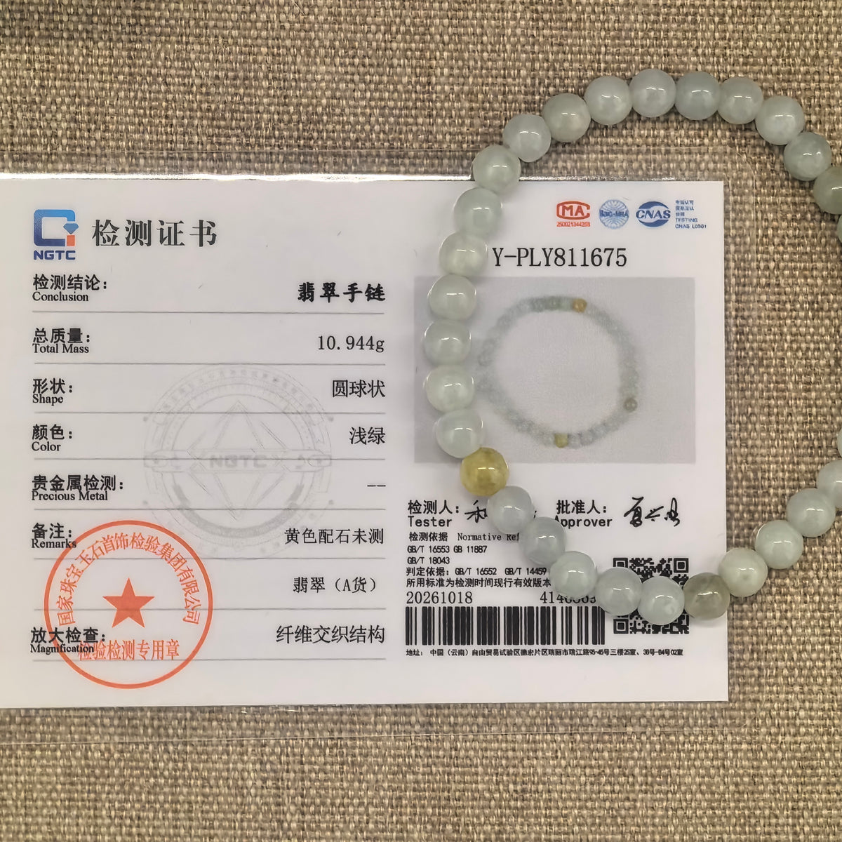 Pale green jadeite bead bracelet with yellow accents beside its matching NGTC certificate on woven fabric.