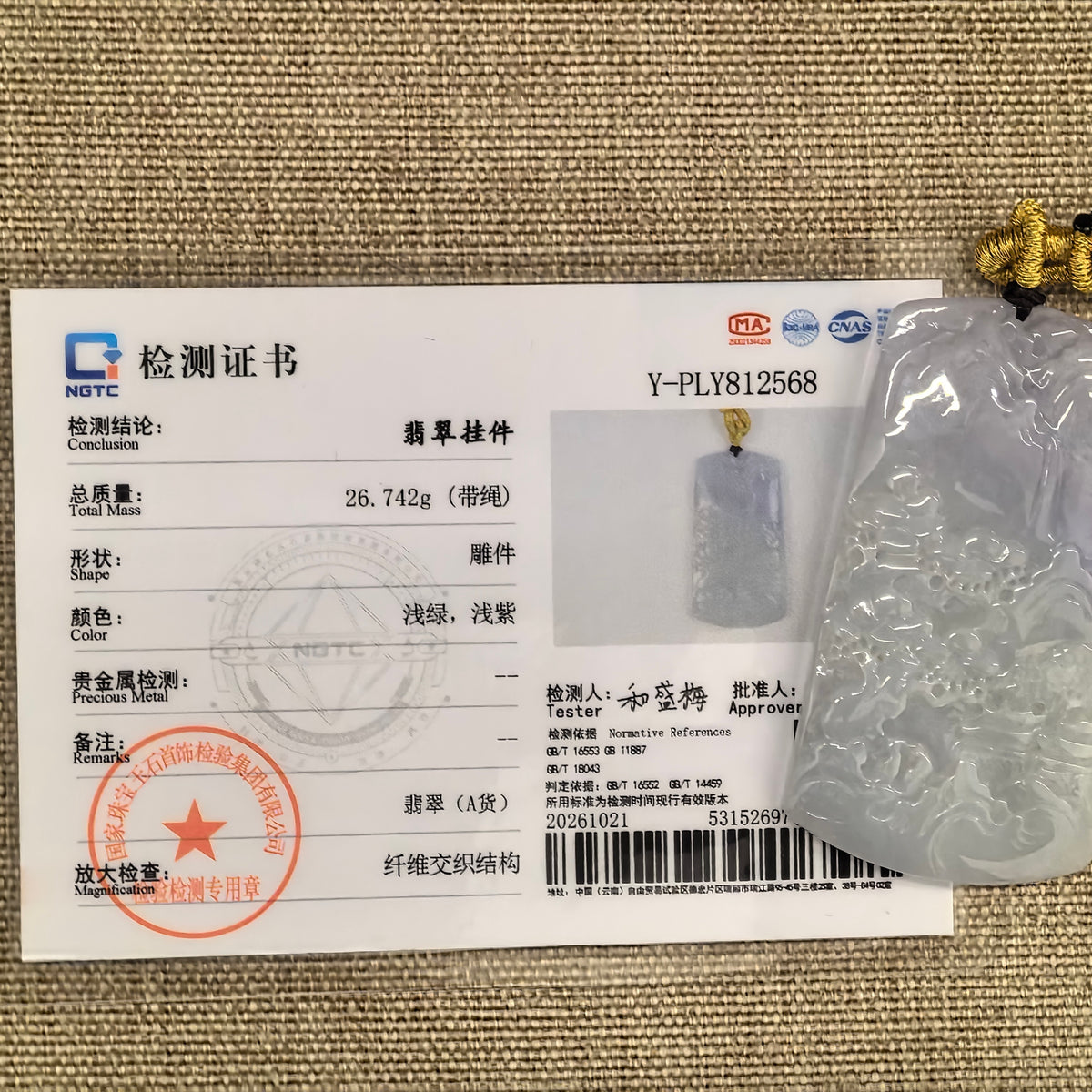 Partly purple jadeite landscape plaque pendant beside its matching NGTC certificate on woven fabric, with its cord and decorative knot visible.