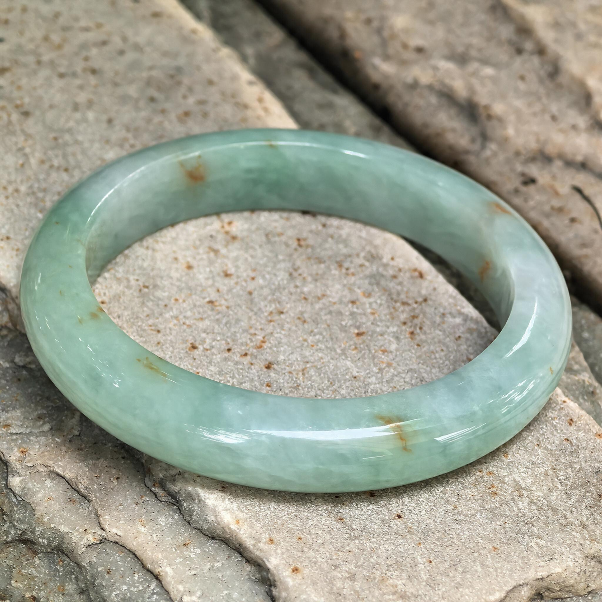 Angled view of the jadeite D-shape bangle on stone, highlighting its softly clouded pale green texture and polished outer surface.