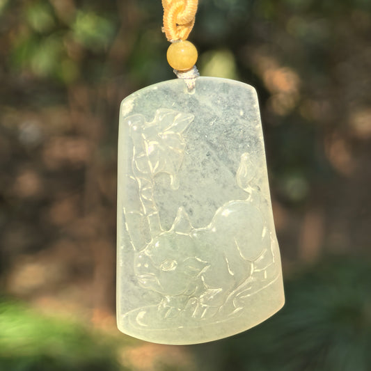 Pale green jadeite Pig Plaque Pendant hanging from a cord with a yellow accent bead, showing its icy translucency and lively carving.