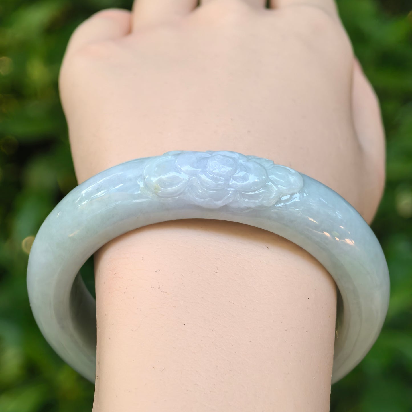 Front view of a light green jadeite oval bangle on the wrist, featuring a delicate carved lotus motif.