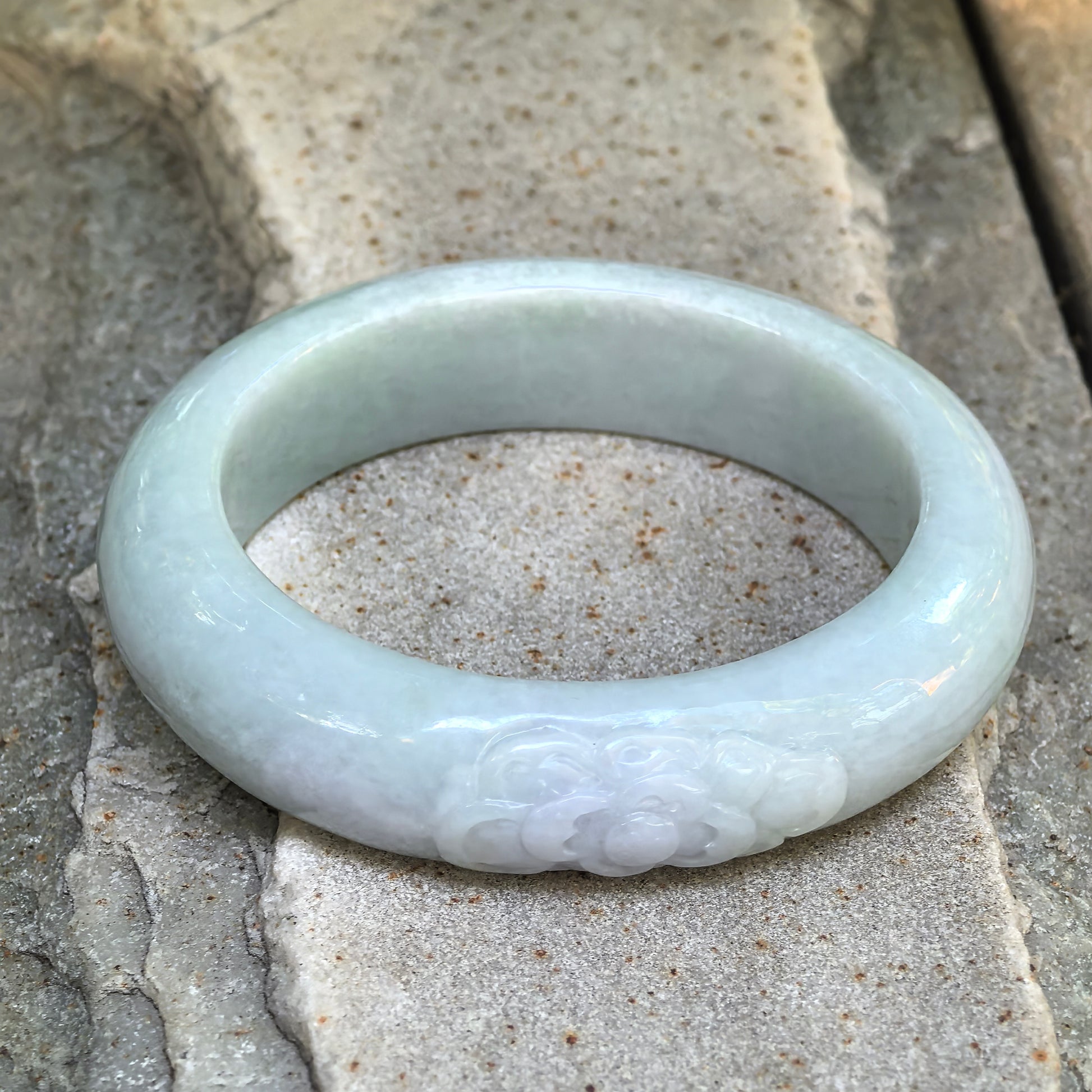 Another full view of the carved lotus jadeite oval bangle on stone, highlighting its light green tone and oval profile.
