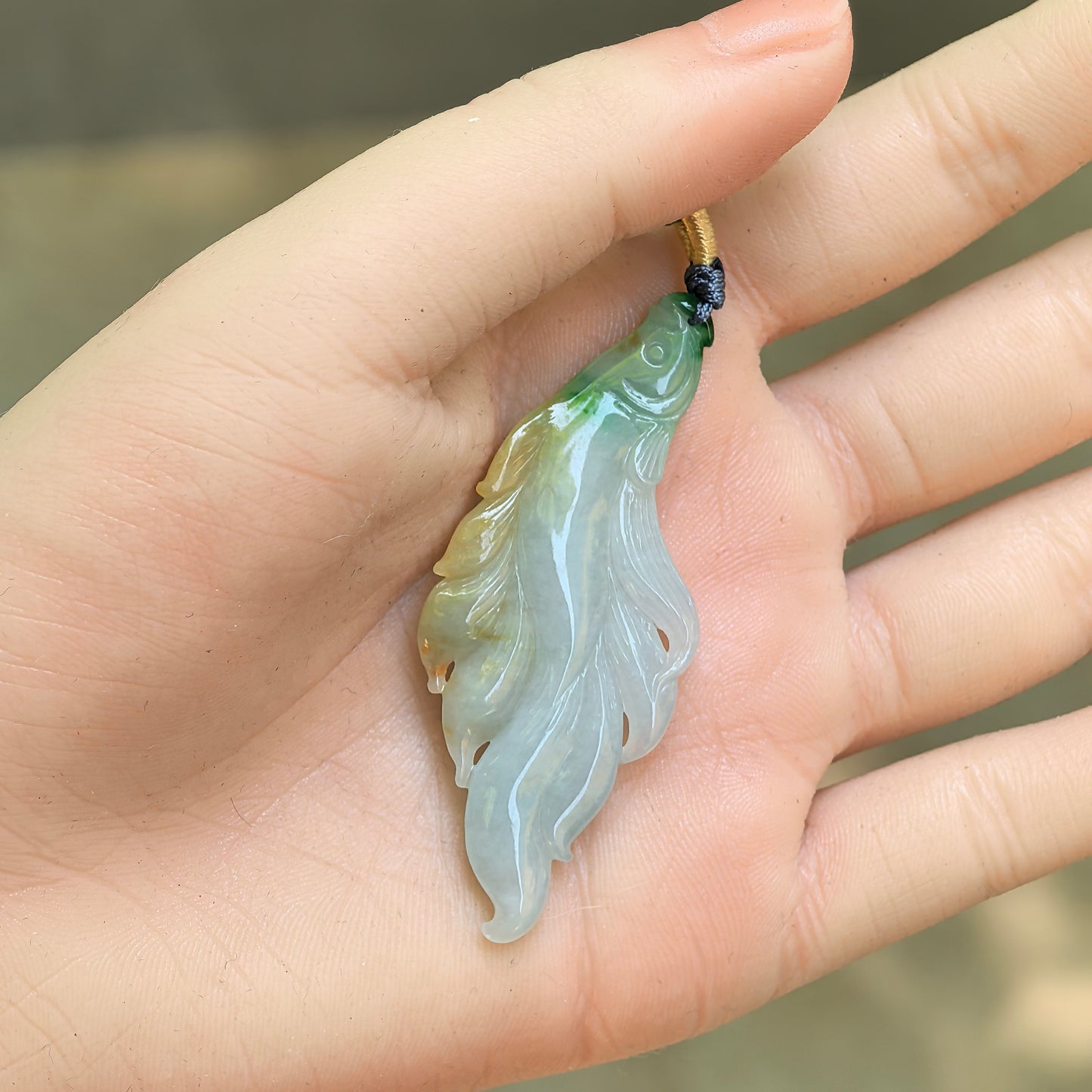 Natural jadeite fish pendant held in hand, showing slender shape and warm yellow-green colour.