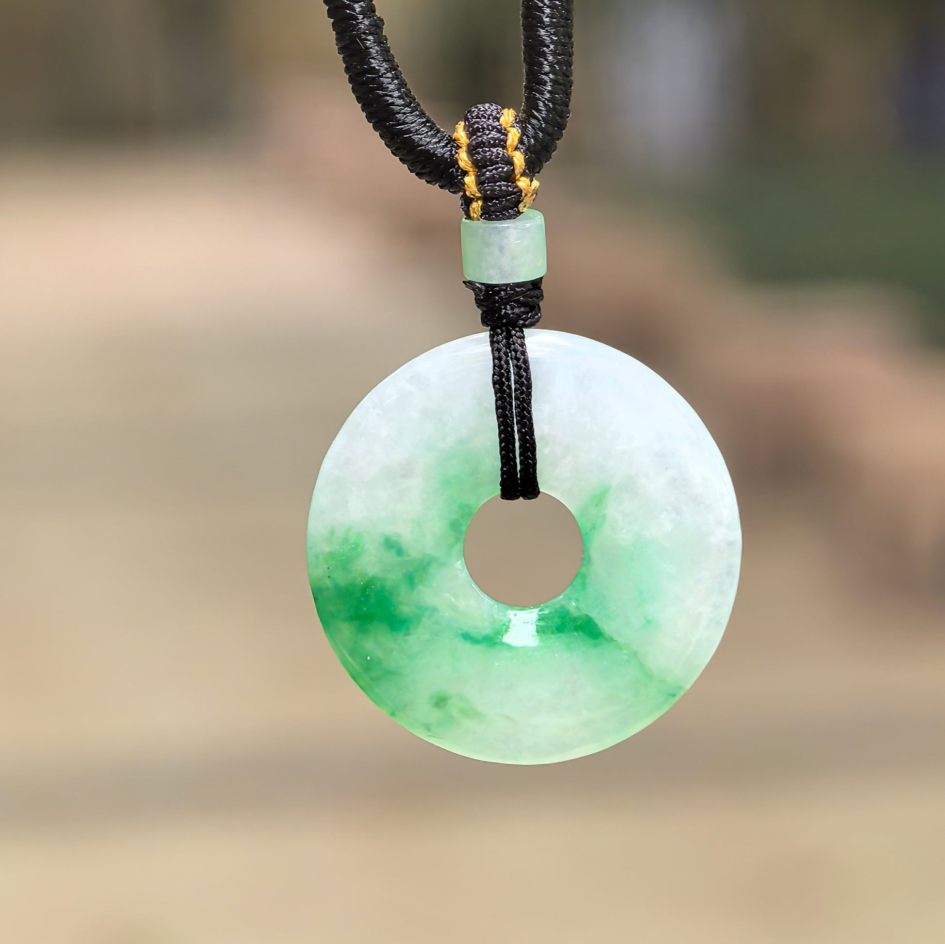 Natural Grade-A glutinous white-based green donut jadeite pendant hanging with black cord against outdoor background.
