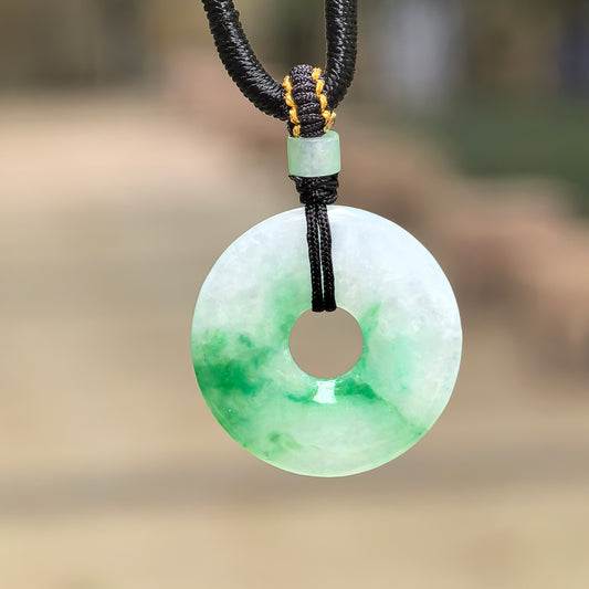 Natural Grade-A glutinous white-based green donut jadeite pendant hanging with black cord against outdoor background.
