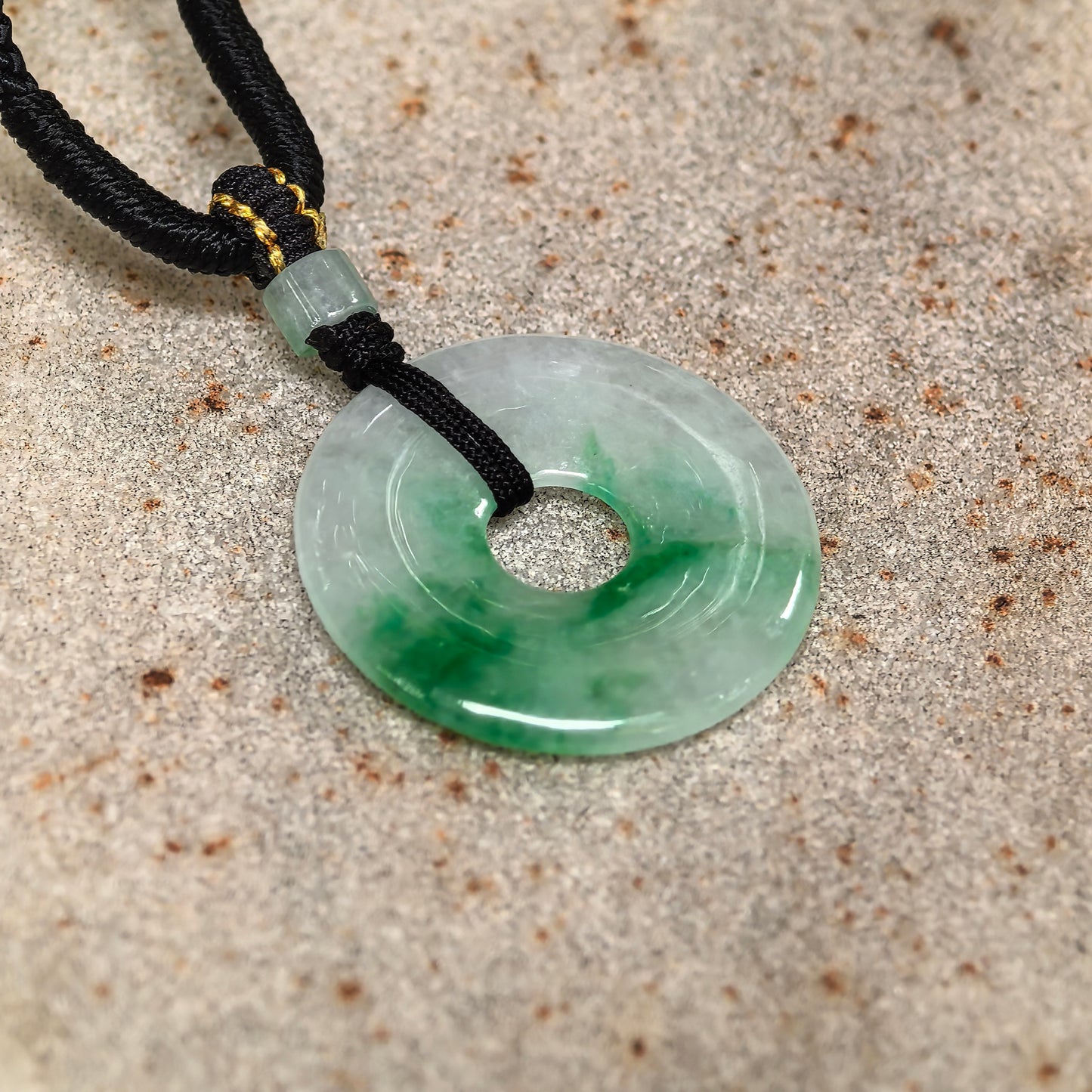 Glutinous white-based green donut jadeite pendant on stone background with cord and small jadeite bead.
