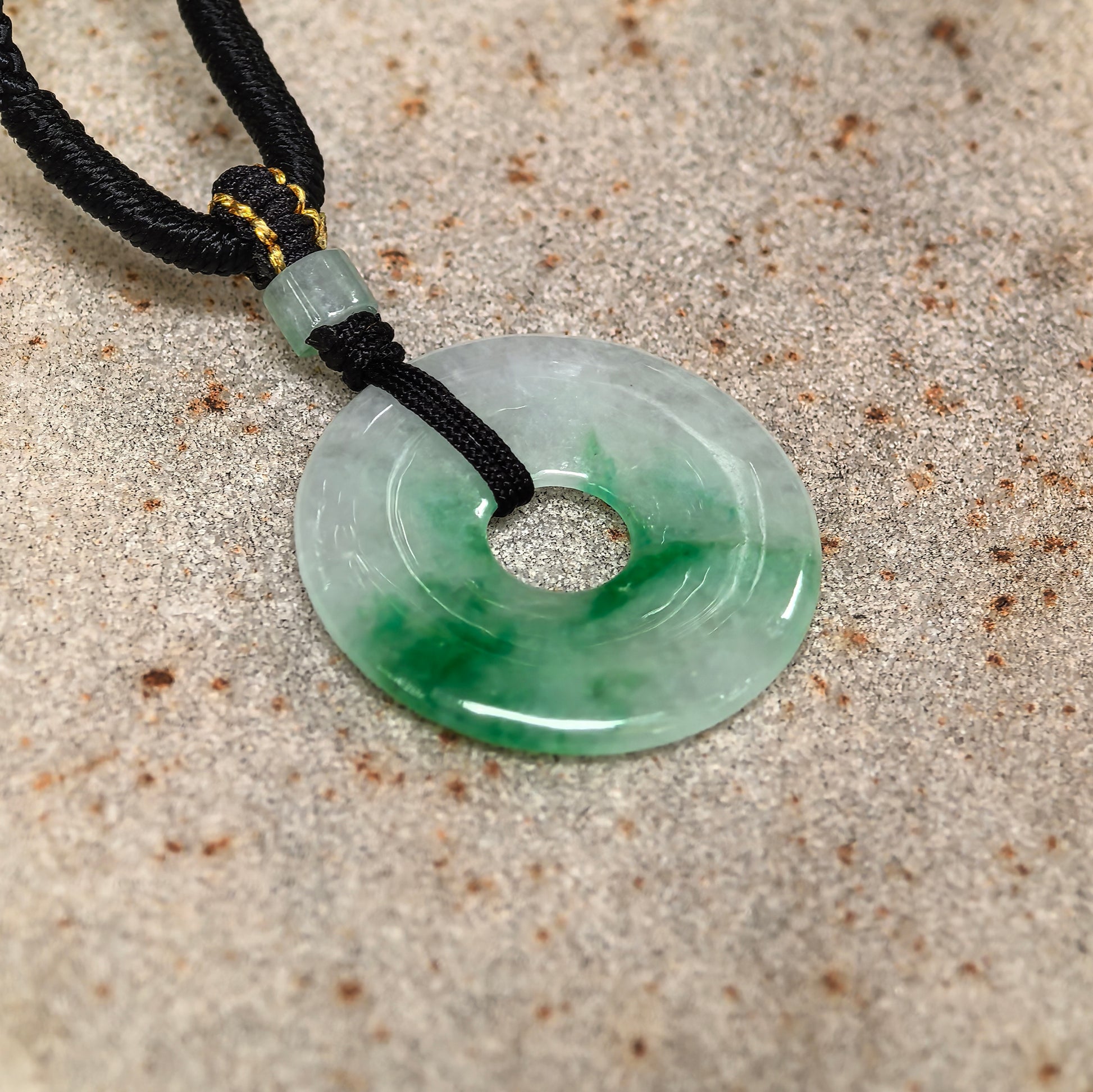 Glutinous white-based green donut jadeite pendant on stone background with cord and small jadeite bead.
