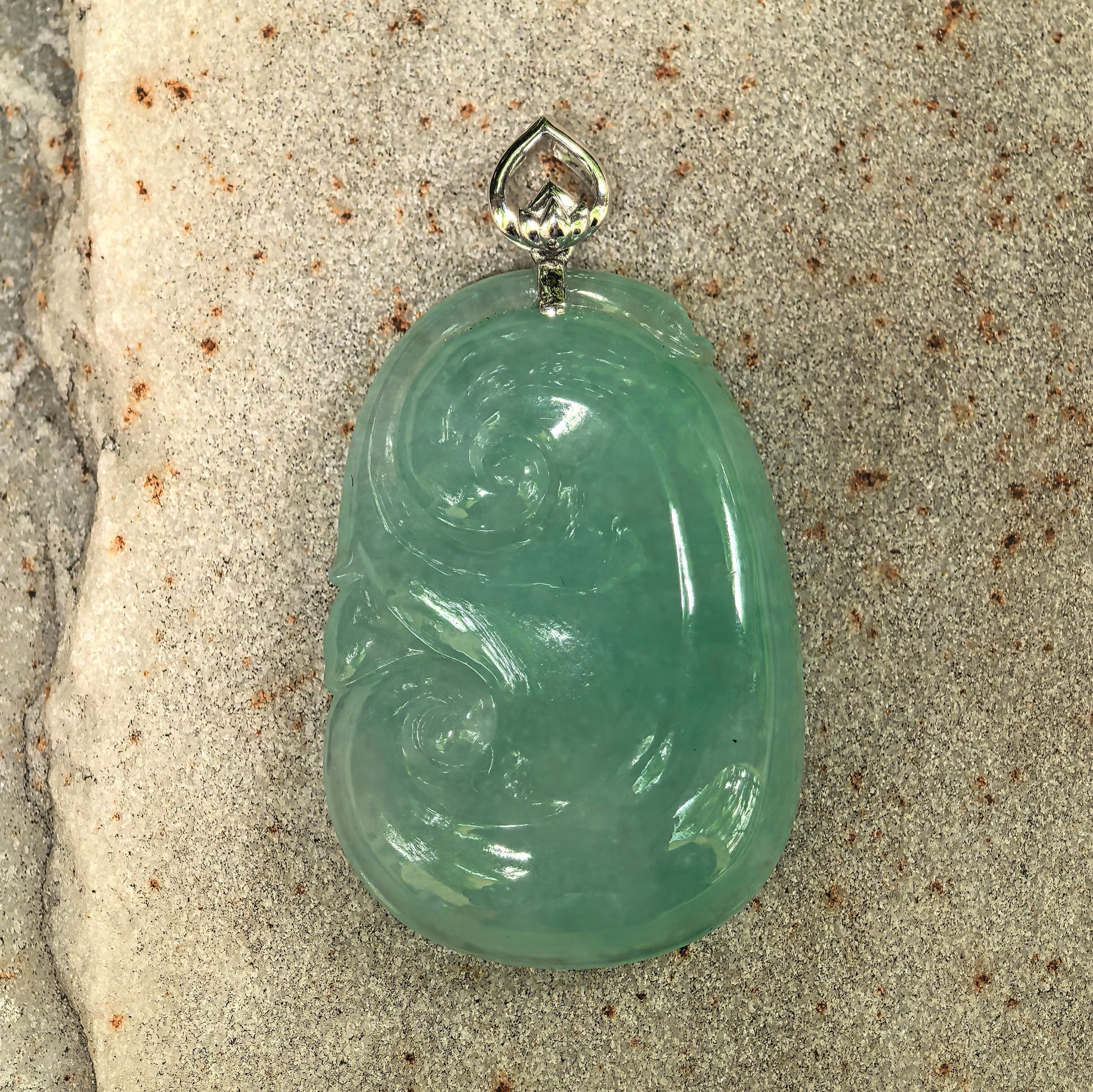 Close-up of icy glutinous fruit green Ruyi jadeite pendant on stone background showing polished surface and carved scroll details.
