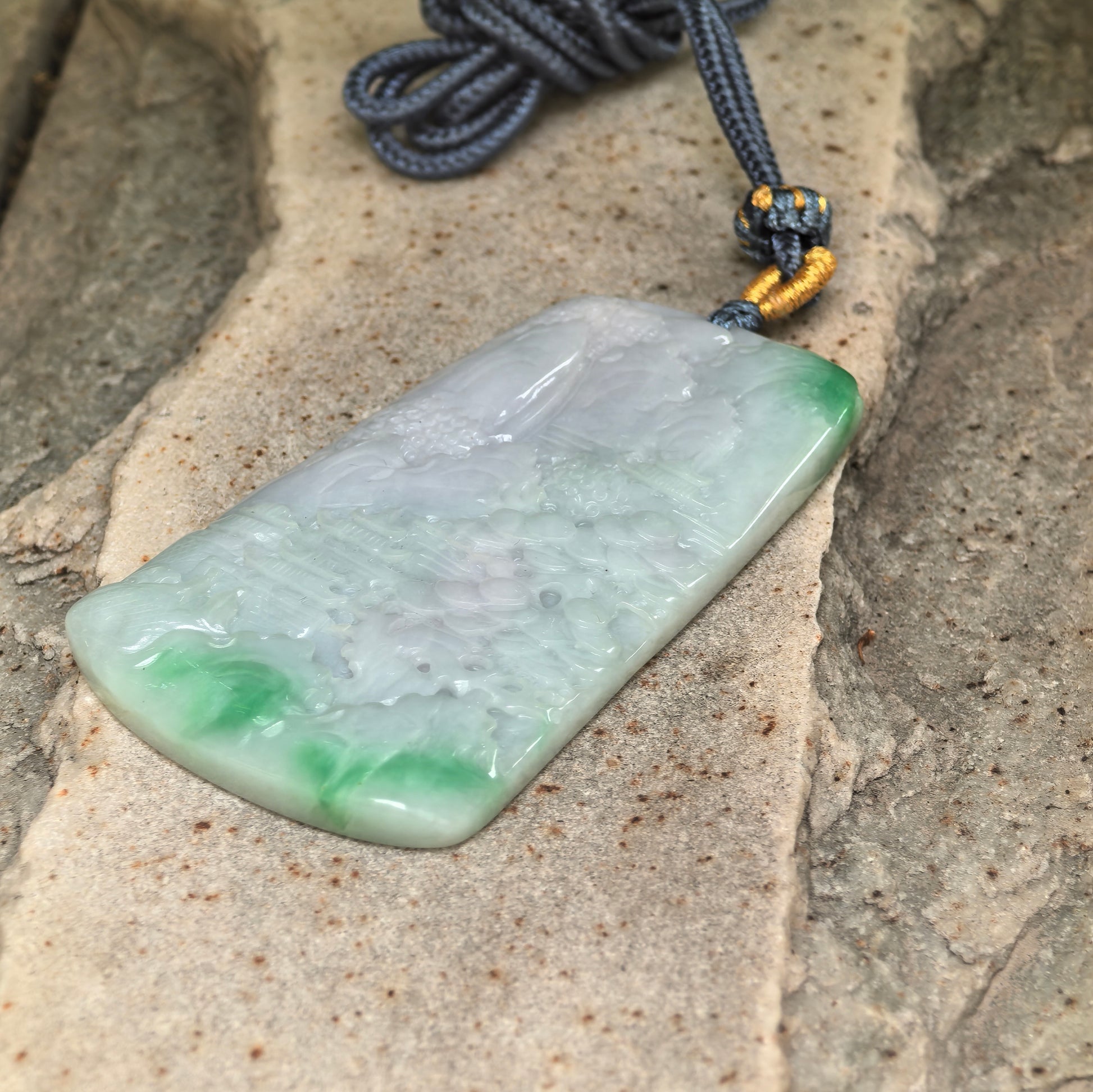 Side view of large white-base green landscape jadeite pendant with cord and polished surface.
