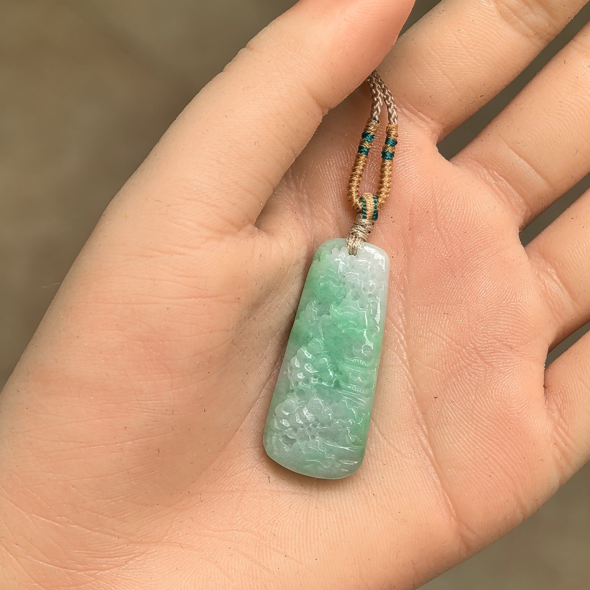 White-base green jadeite landscape pendant shown in hand with slim plaque shape and cord.
