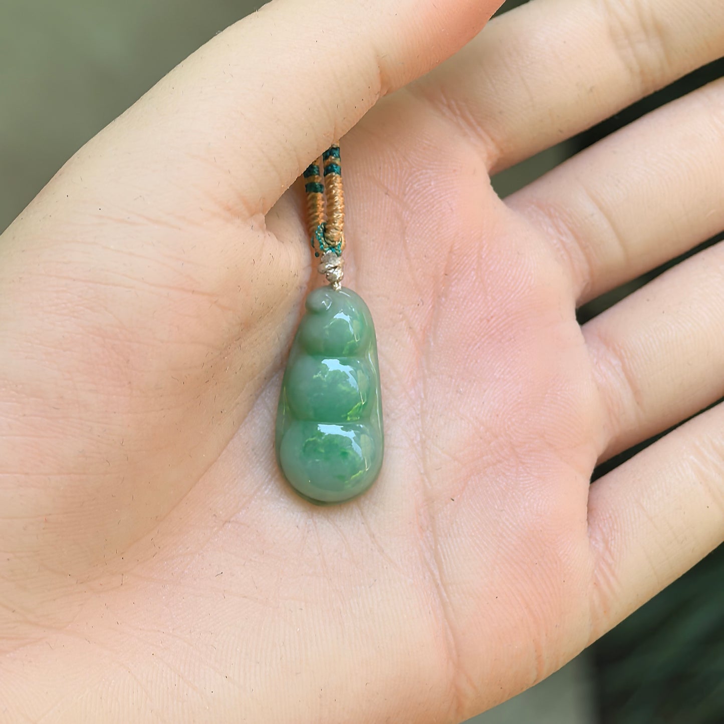Small green jadeite bean pendant displayed in hand, showing rounded three-bean carving and smooth polish.
