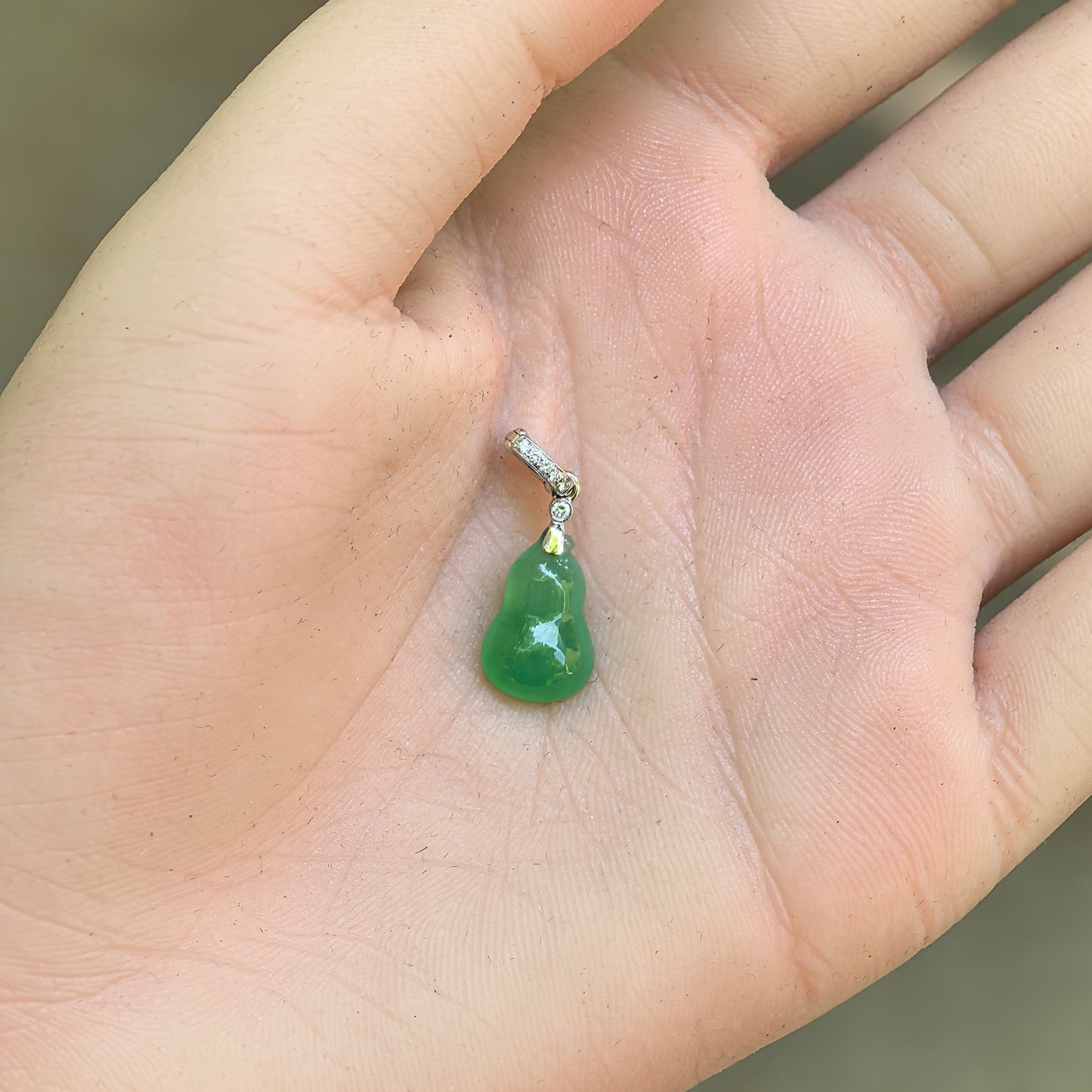 Small vivid green gourd jadeite pendant shown on hand with synthetic diamond accented bail.
