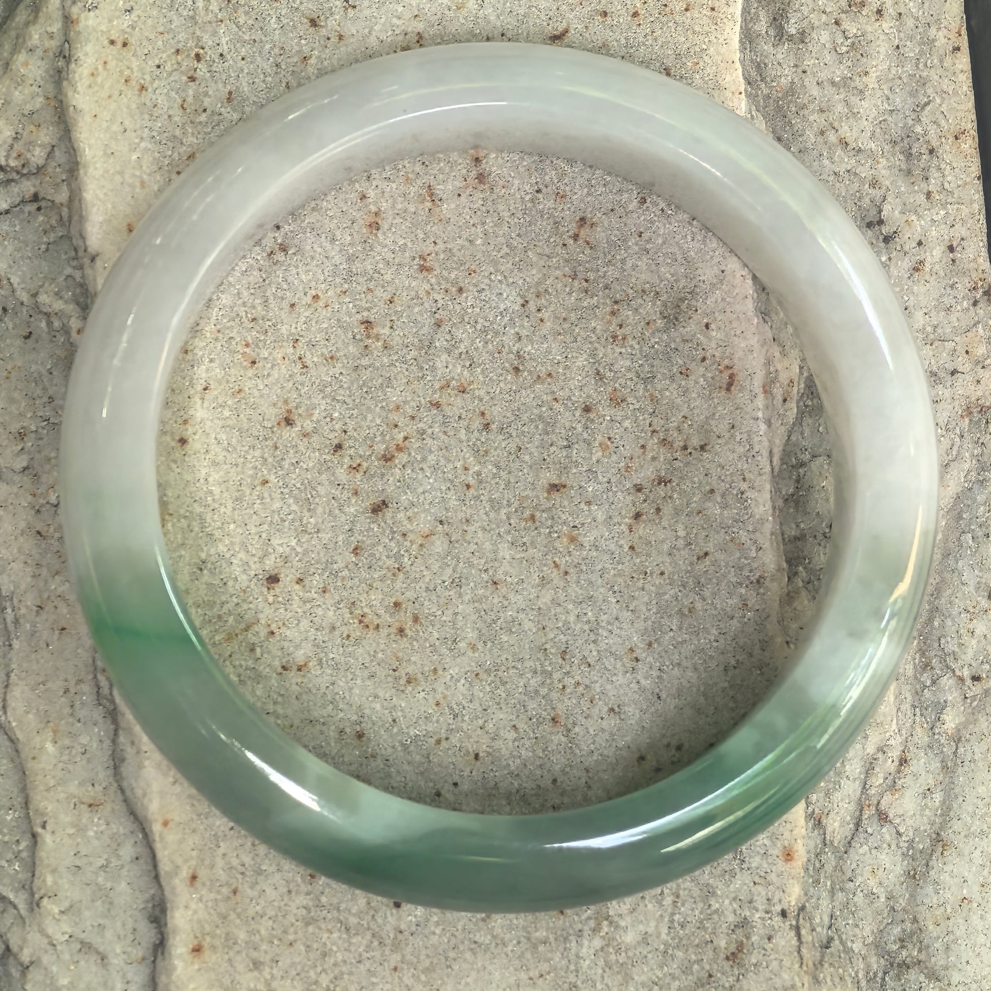 Fine glutinous Burmese jadeite bangle with soft white base and green sections on stone background.
