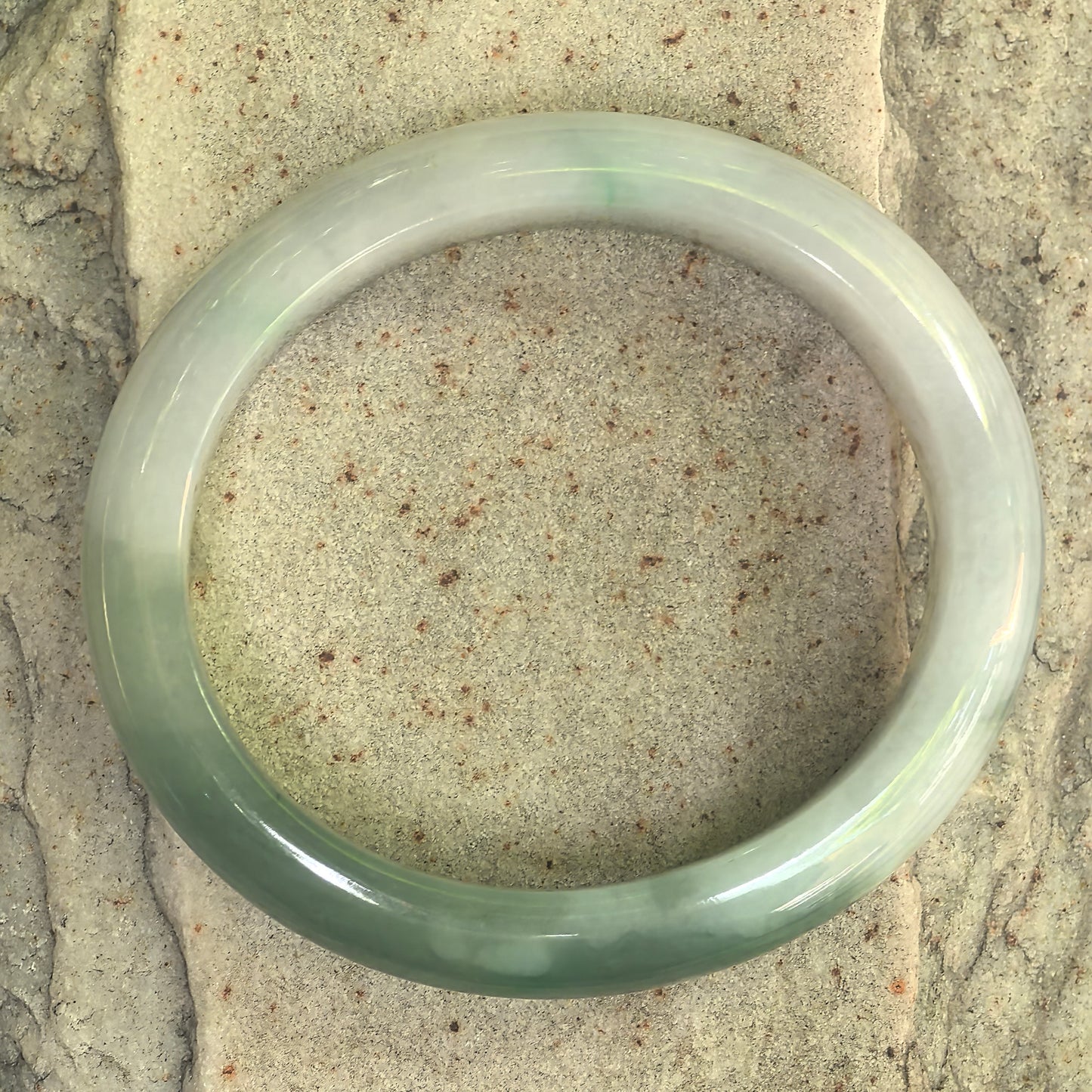 Top view of glutinous white and light green oval jadeite bangle displayed on stone surface.
