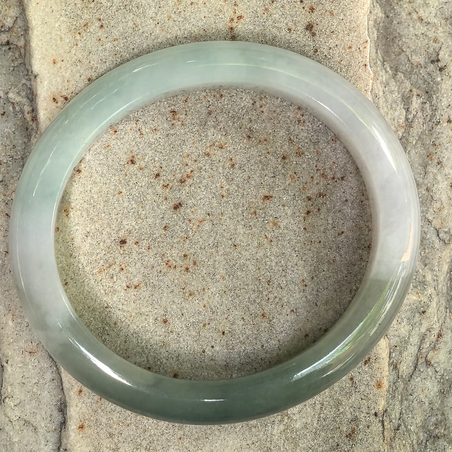 White-base jadeite oval bangle with dark green areas displayed flat on a stone background.
