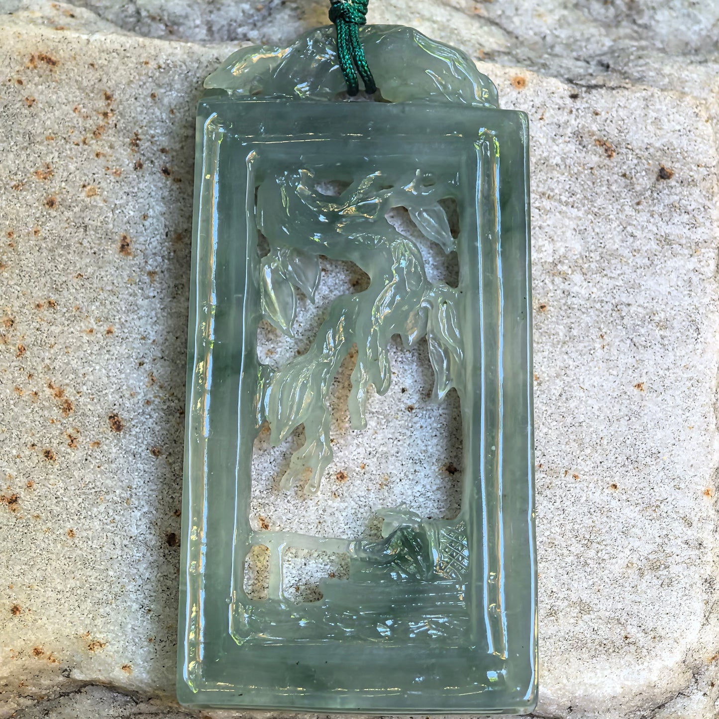 Close-up of icy glutinous pale green jadeite pendant showing hollowed-out fishing landscape details.
