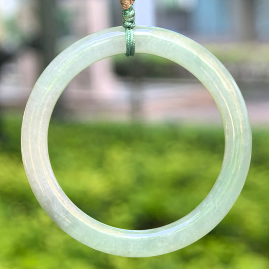 Natural Grade-A glutinous light green child jadeite bangle hanging with cord against a soft green background.
