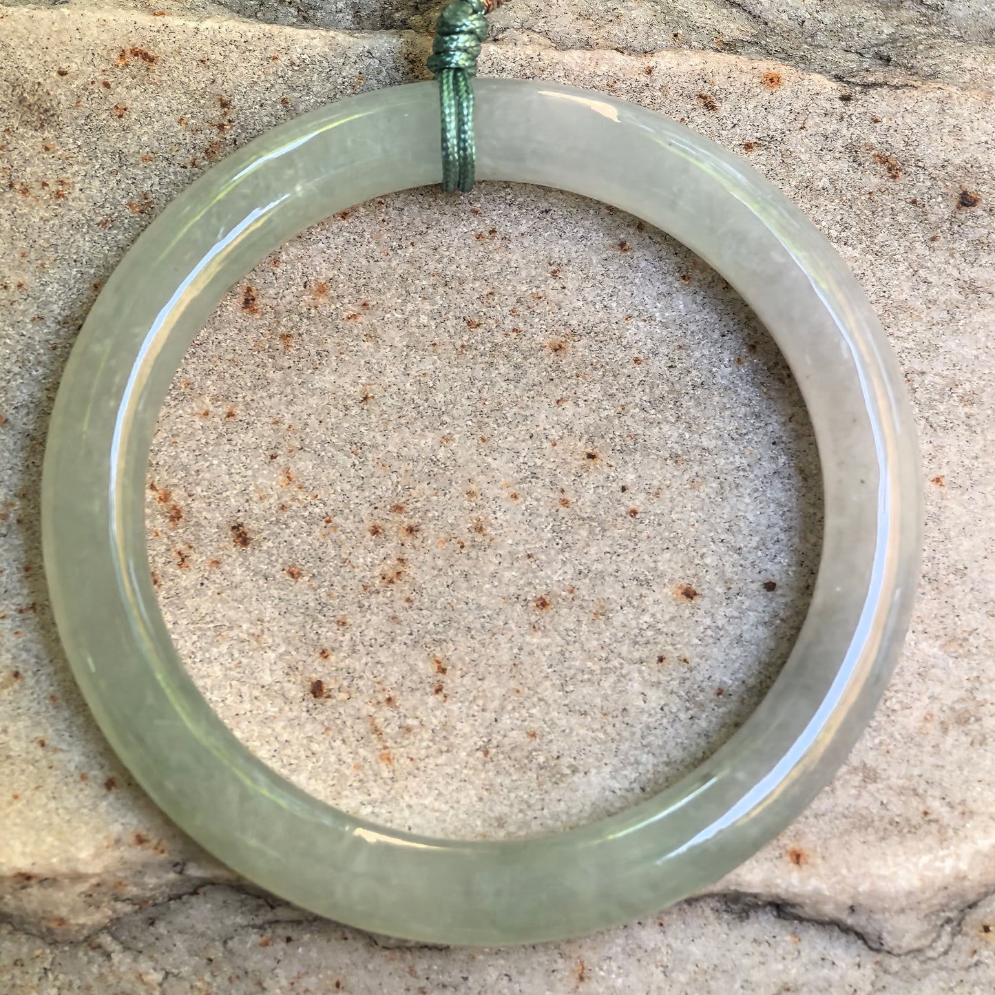 Glutinous light green jadeite bangle displayed flat on stone with cord, suitable as a small bangle or pendant.
