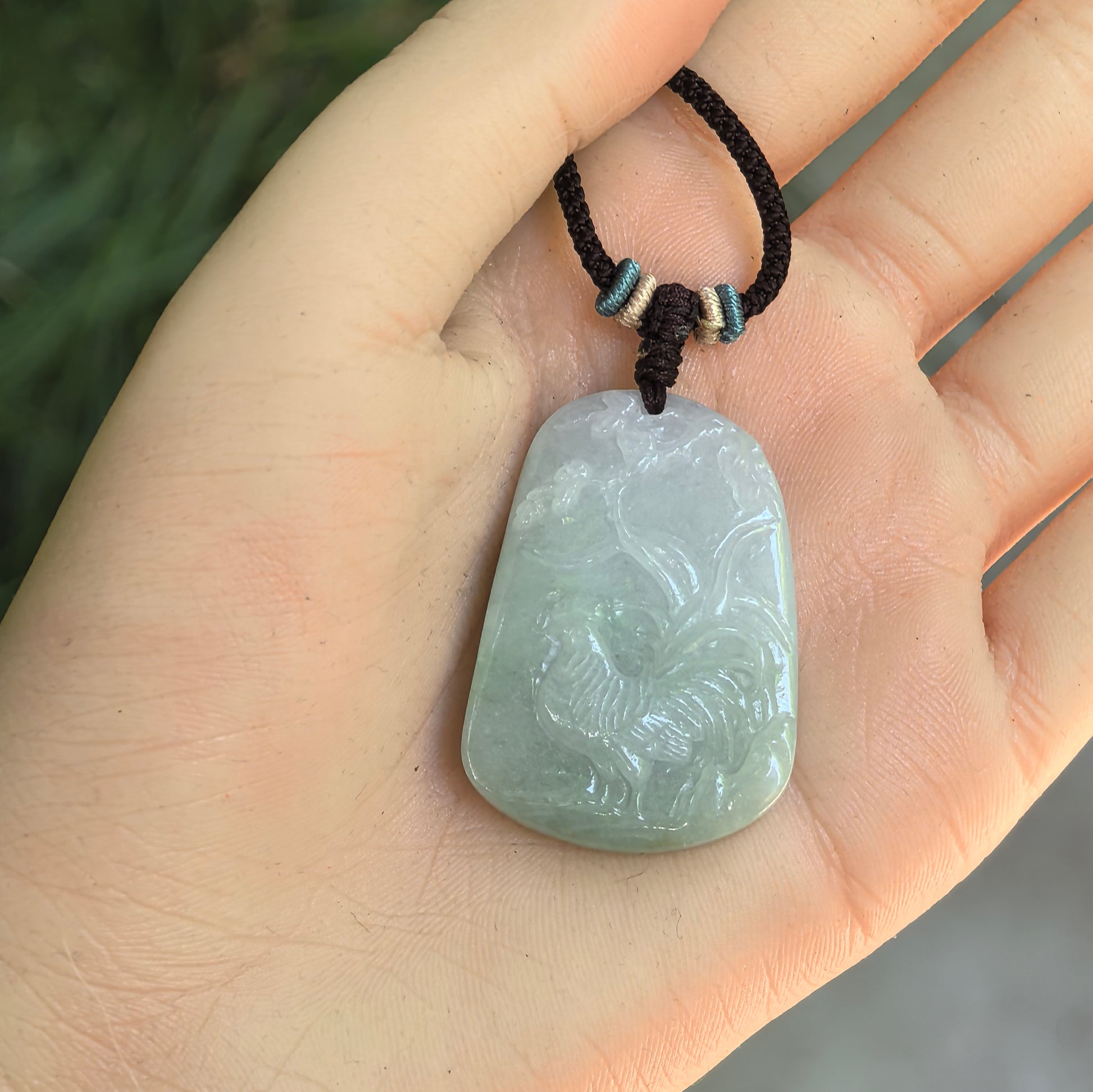 Glutinous light green jadeite rooster pendant displayed in hand with rounded plaque-like shape.
