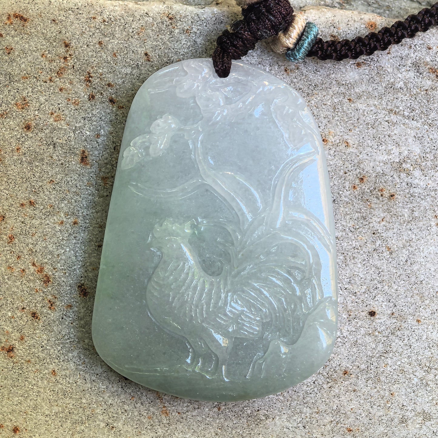 Natural Grade-A light green rooster jadeite pendant shown on stone surface with floral branch carving.
