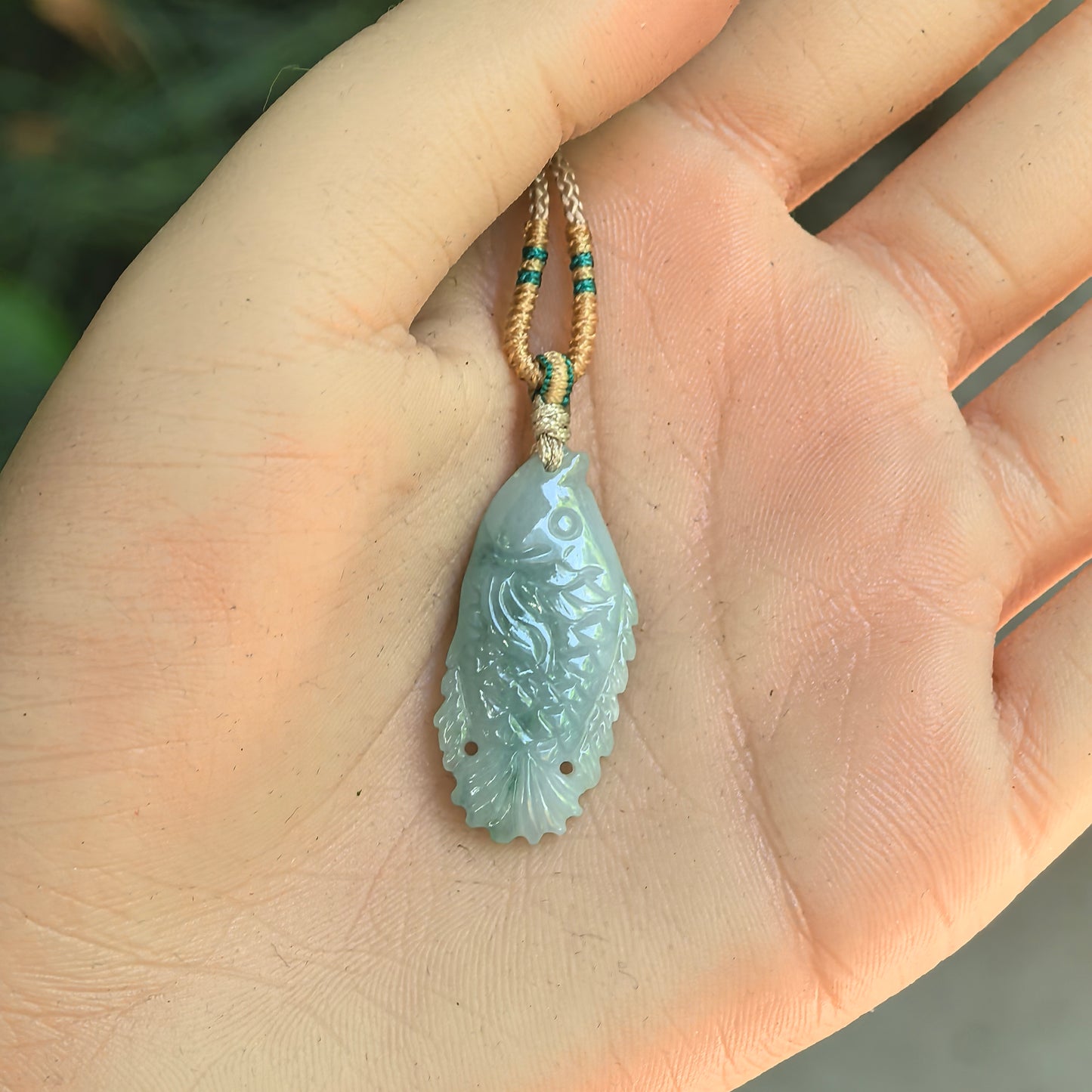 Glutinous light green fish jadeite pendant displayed in hand with slim carved fish shape.

