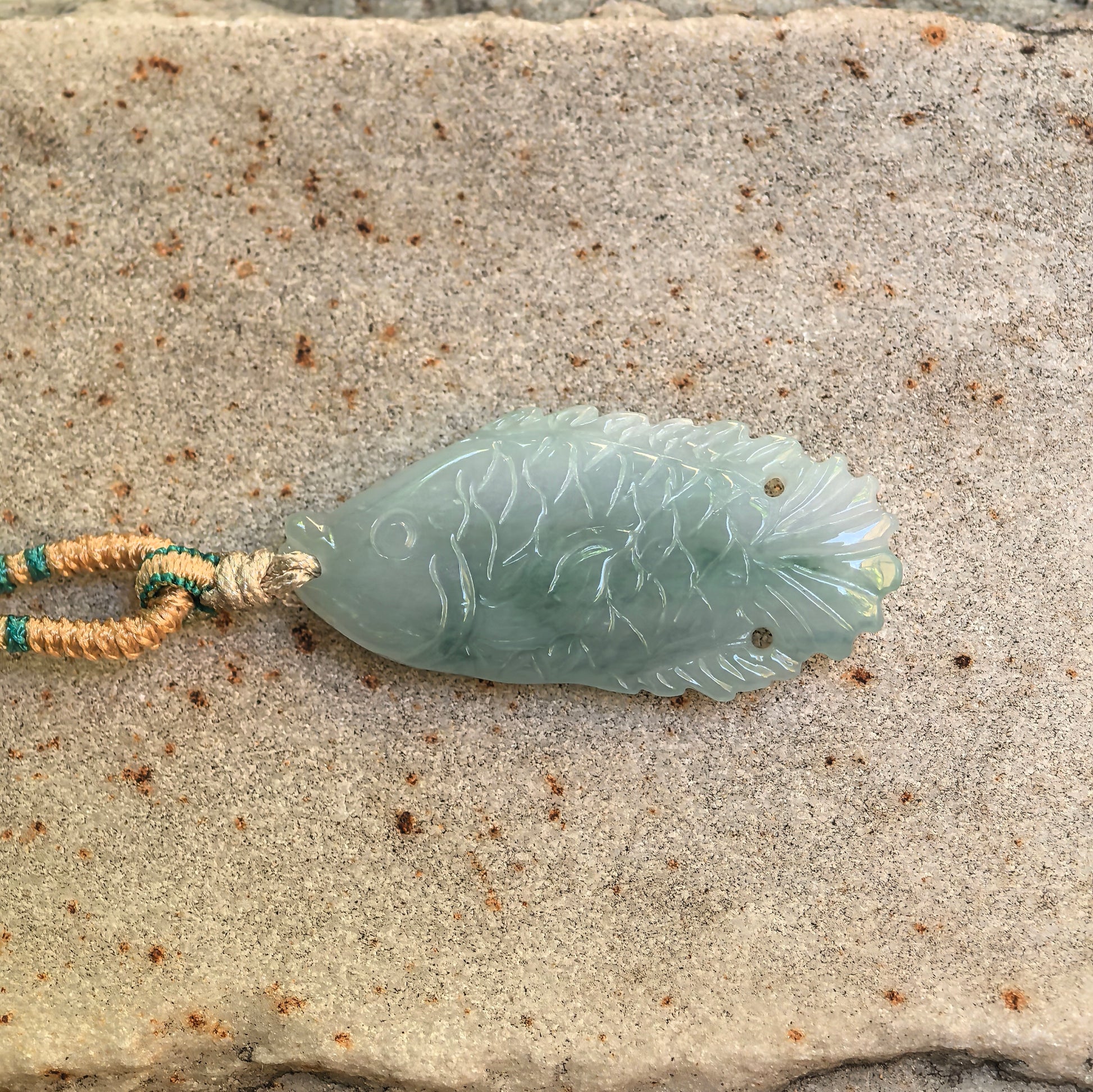Natural Grade-A light green fish jadeite pendant shown on stone surface with carved scale and tail details.
