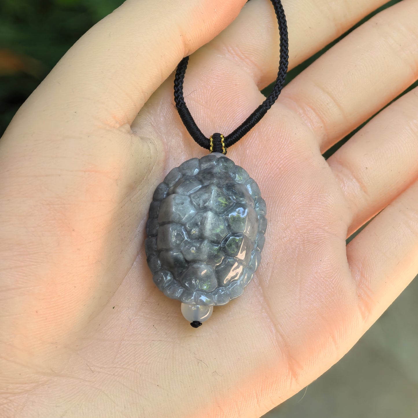 Glutinous gray-black turtle shell jadeite pendant displayed in hand with cord and small bead accent.
