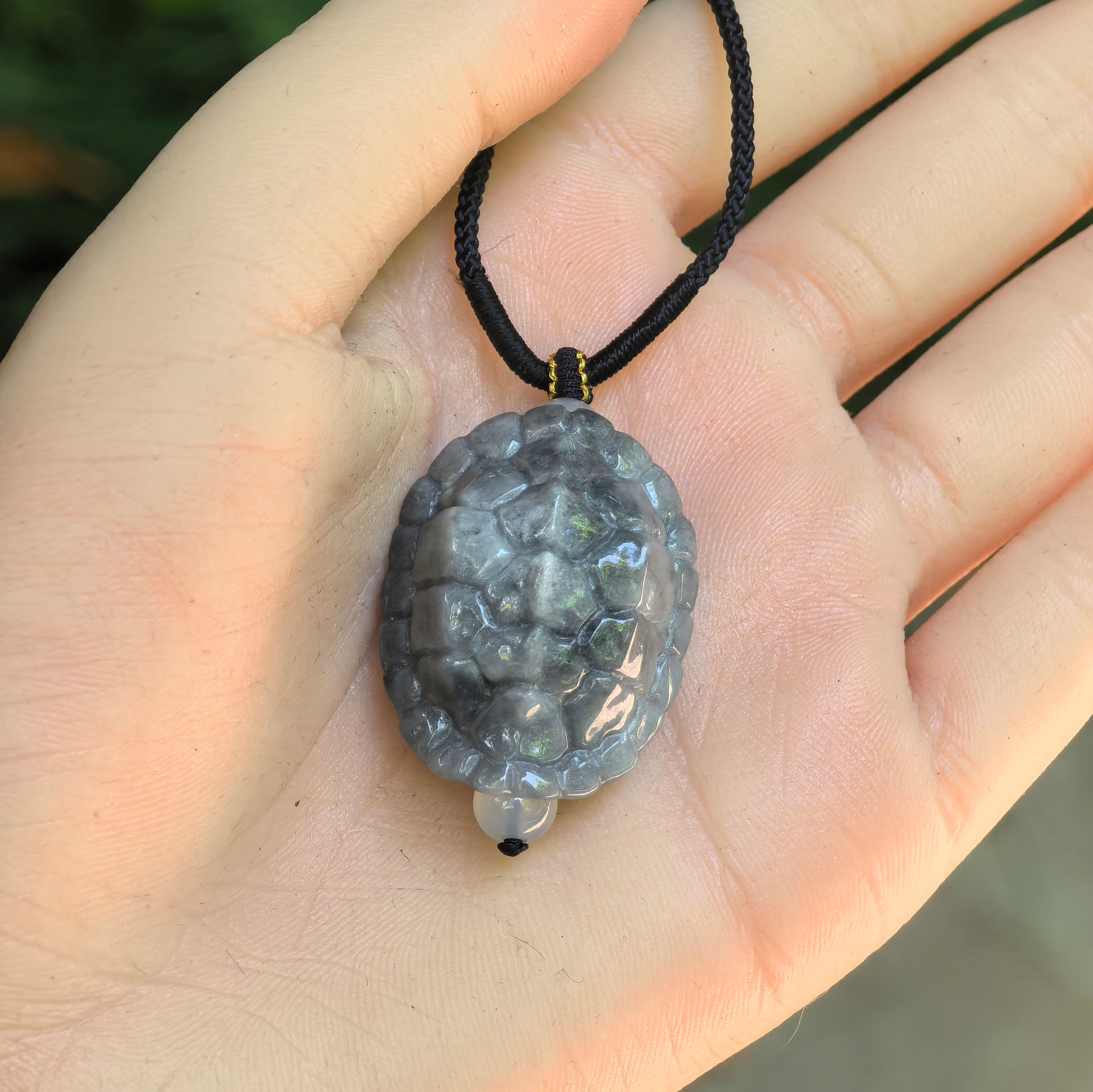 Glutinous gray-black turtle shell jadeite pendant displayed in hand with cord and small bead accent.

