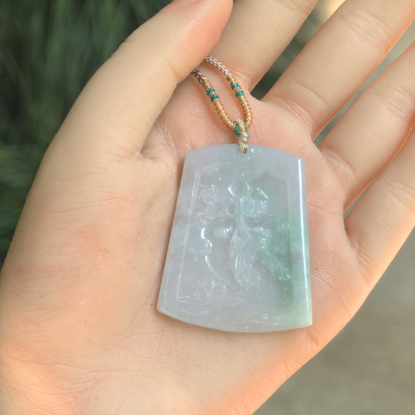 Glutinous white-green jadeite floral plaque pendant shown on hand with cord attached.
