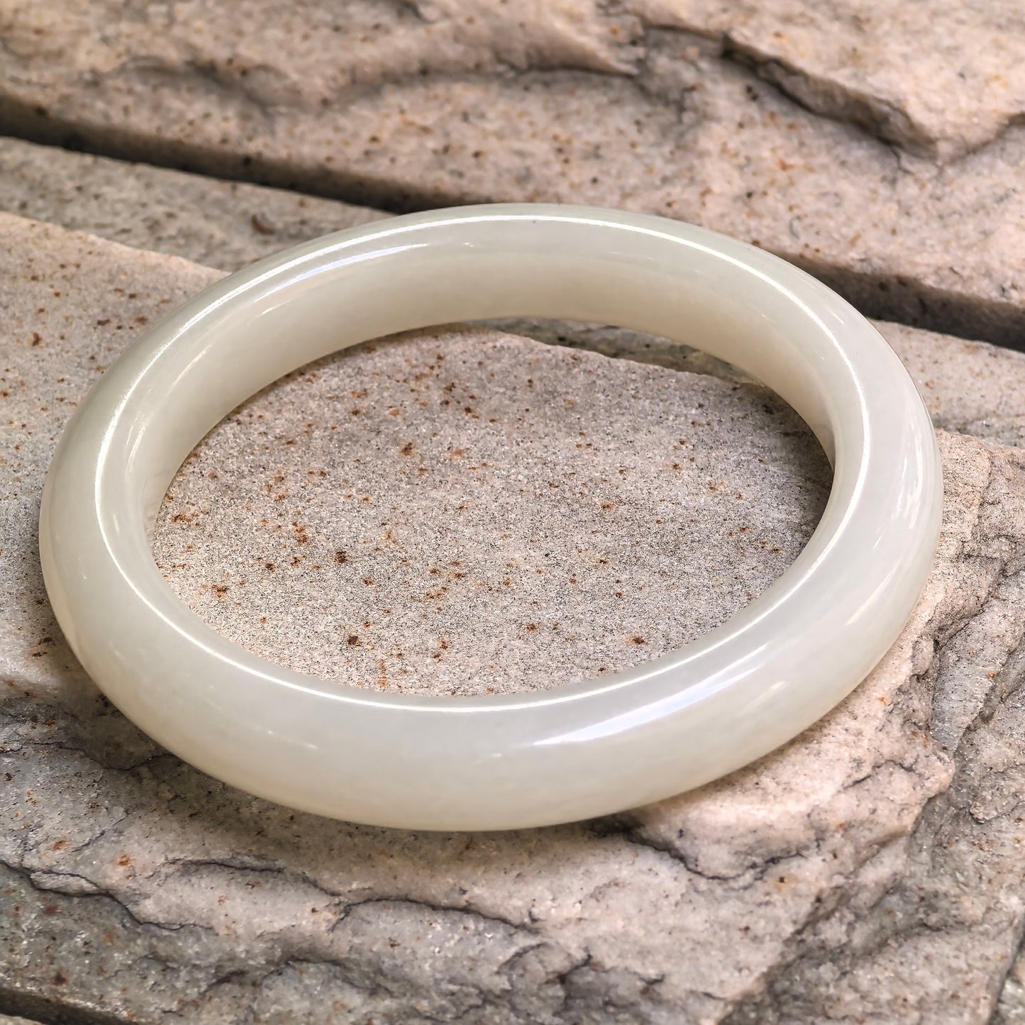 Angled view of natural Hetian jade bangle on stone surface, highlighting its smooth polish and rounded bracelet form.