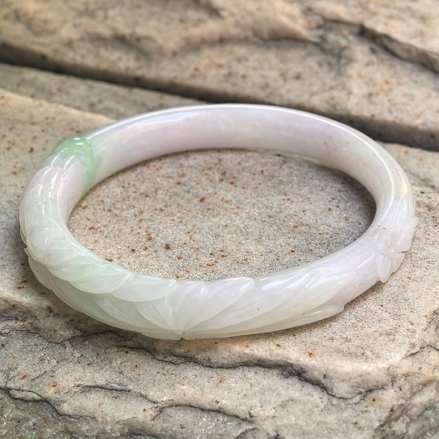 Angled view of Natural Grade-A glutinous floral oval jadeite bangle resting on stone background.
