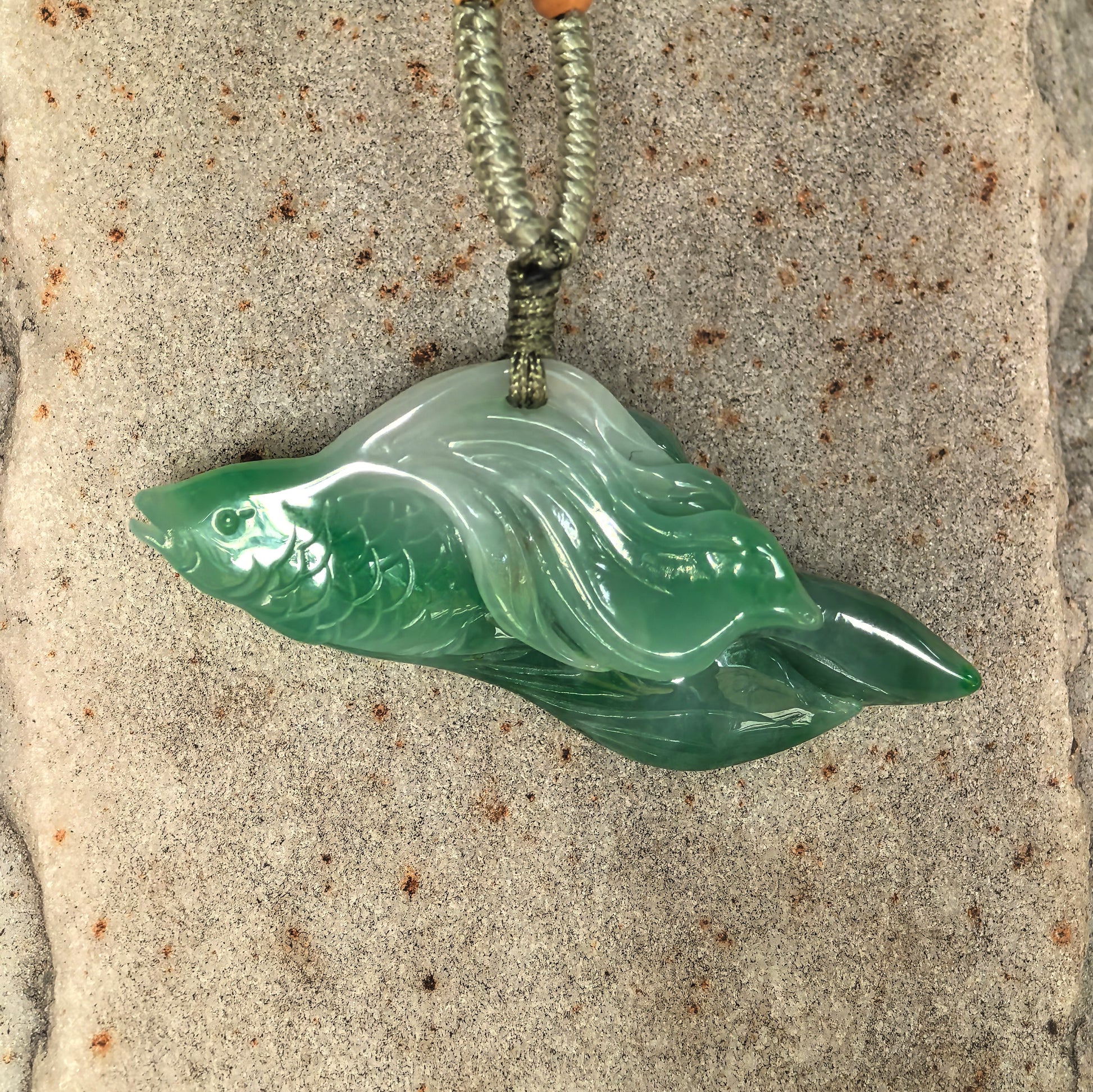 Three-dimensional green jadeite fish pendant displayed on stone surface with detailed scales, fins, and flowing tail.