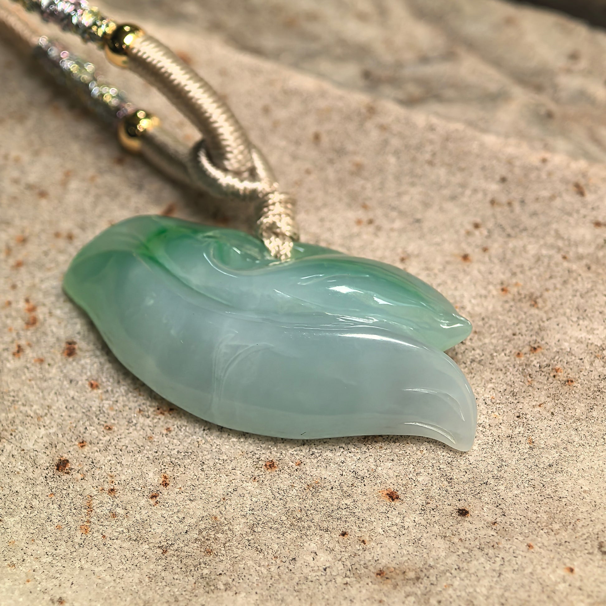 Green and light green jadeite dolphin pendant displayed on stone surface with flowing carved wave lines and polished finish.