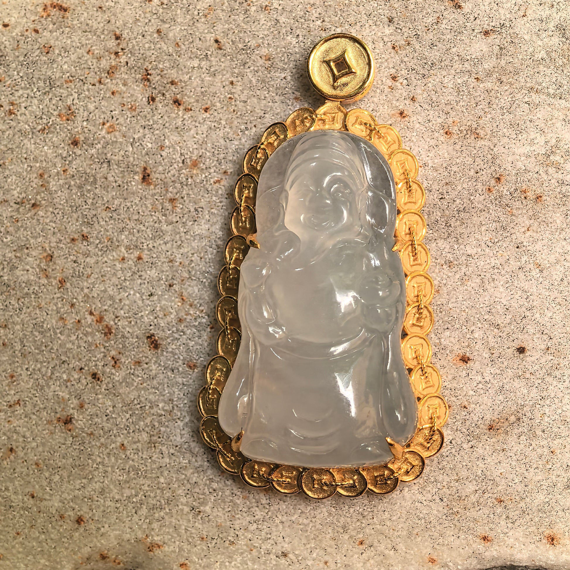 Natural jadeite God of Wealth pendant displayed on stone surface with warm gold setting.