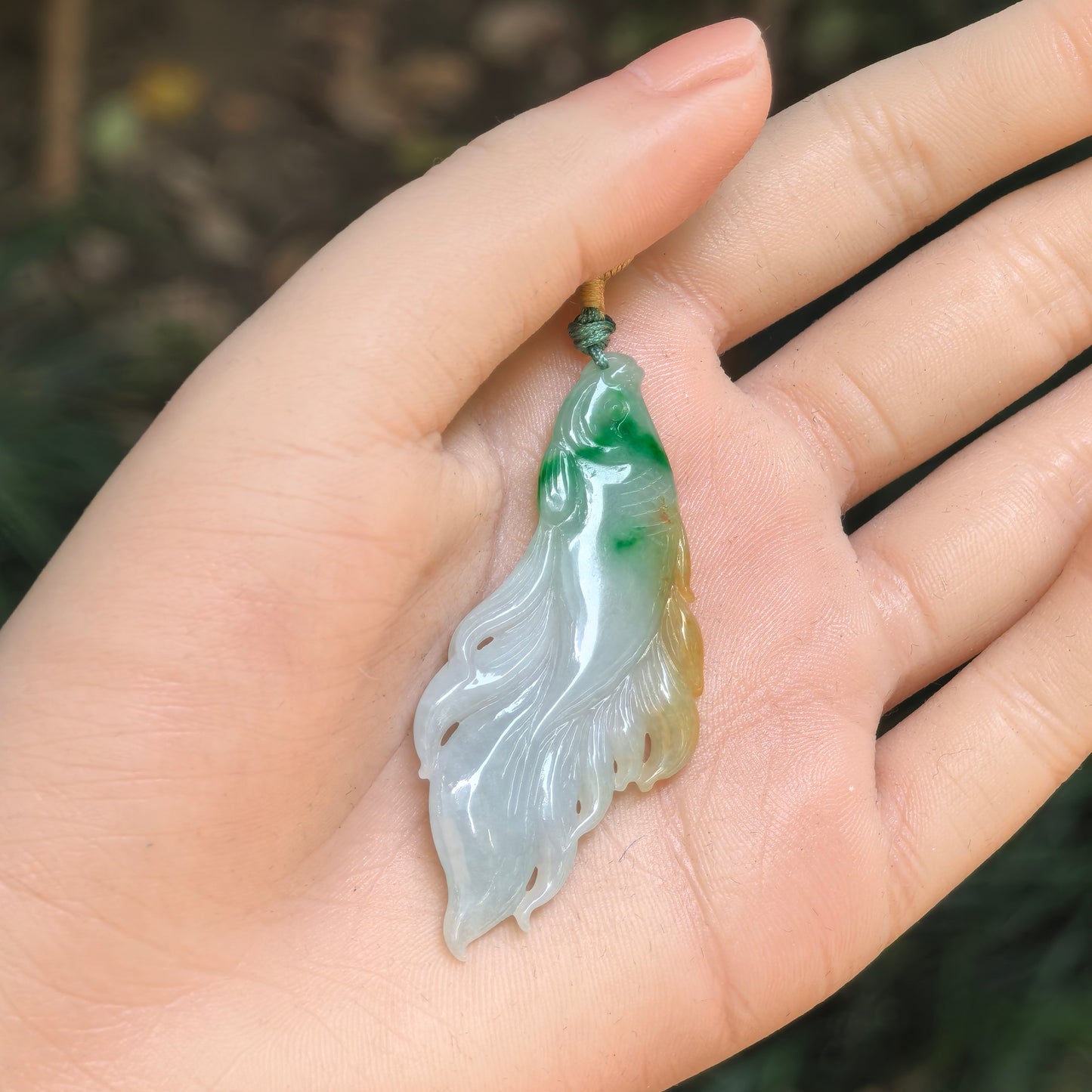 Natural jadeite fish pendant held in hand, showing slender shape and warm yellow-green colour.